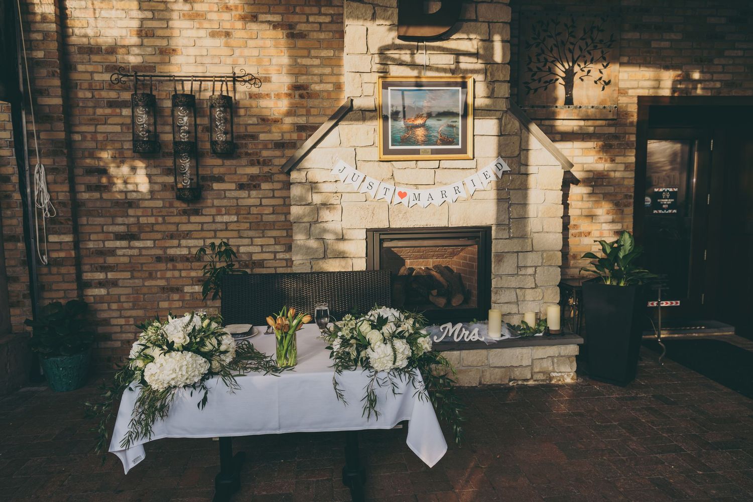 A floral arrangement on a white tablecloth sits in front of a rustic exposed brick wall with fireplace and framed artwork.