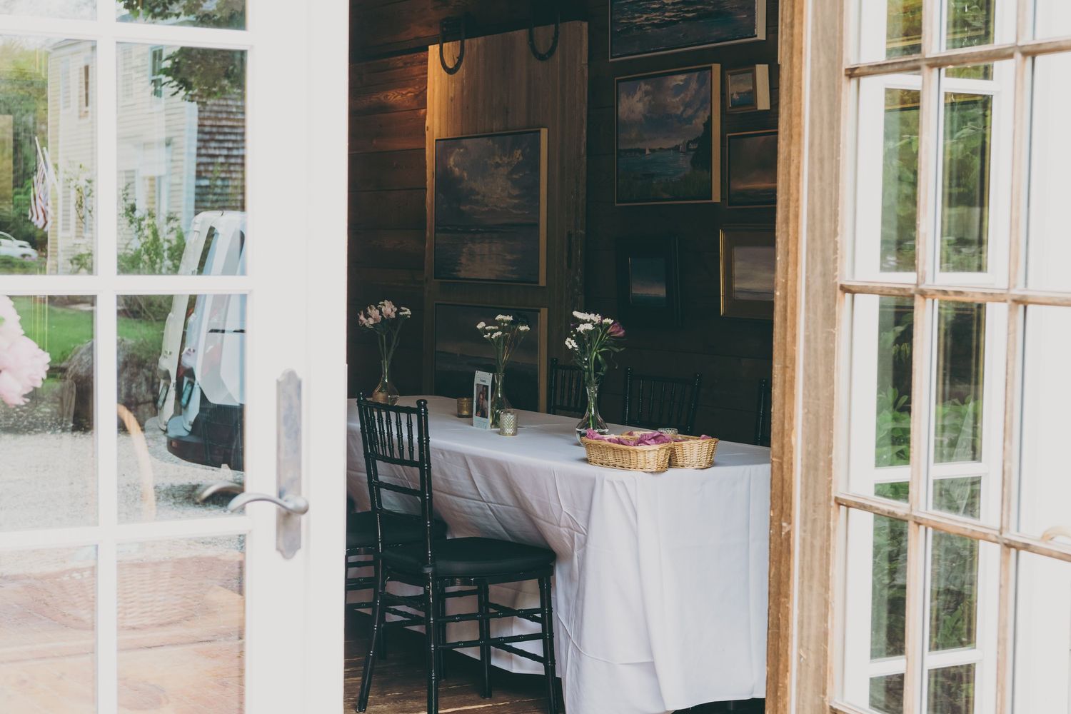 Sunlit dining room interior with white set tables, rustic wooden beams, and large windows overlooking outdoor greenery.