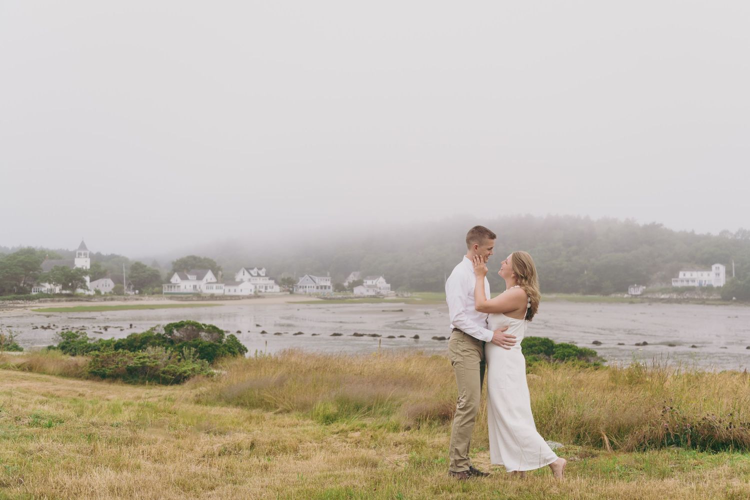 A couple embraces on a grassy hillside overlooking a misty coastal inlet with houses in the background.