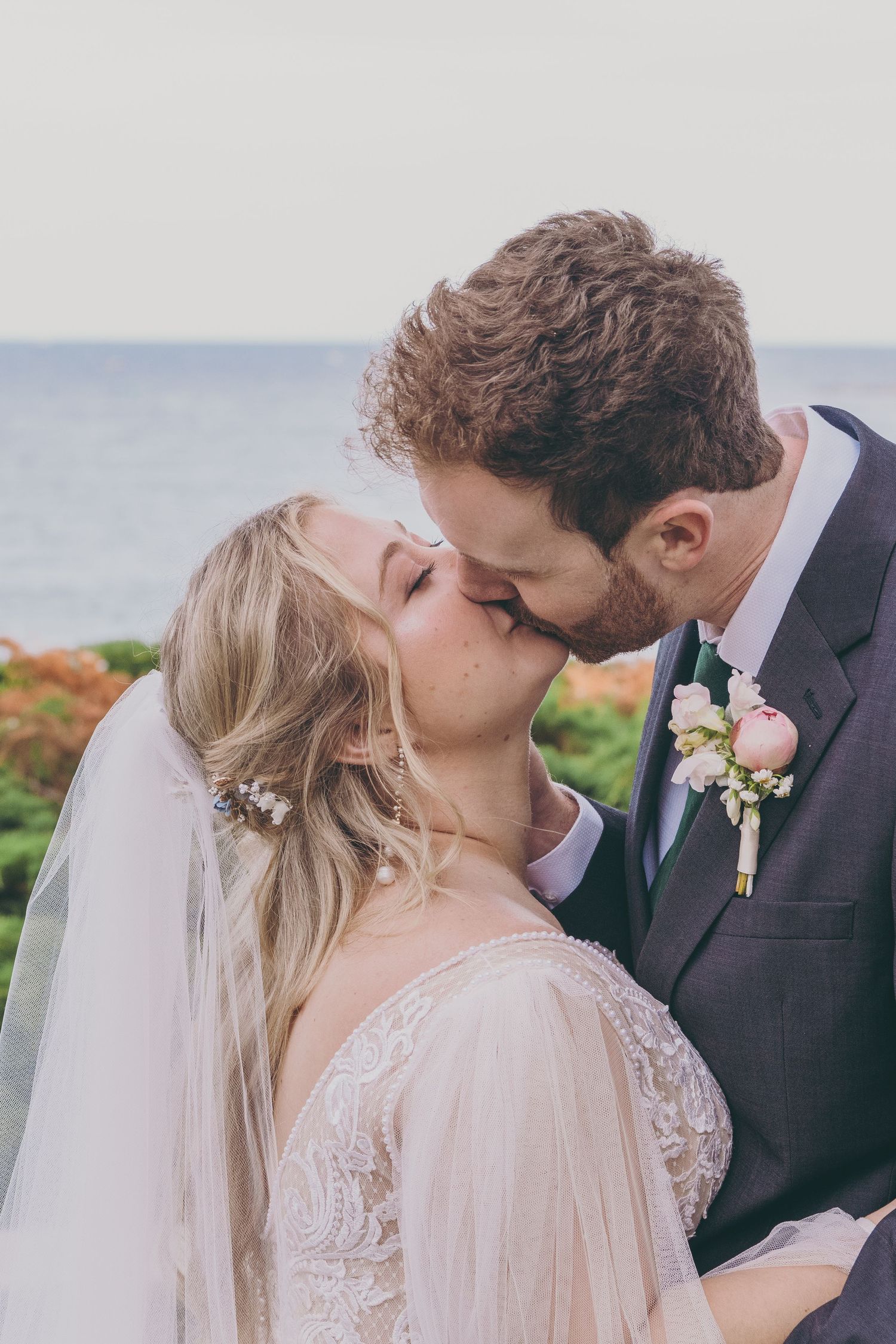 Romantic wedding kiss with bride in lace dress and veil by the ocean.