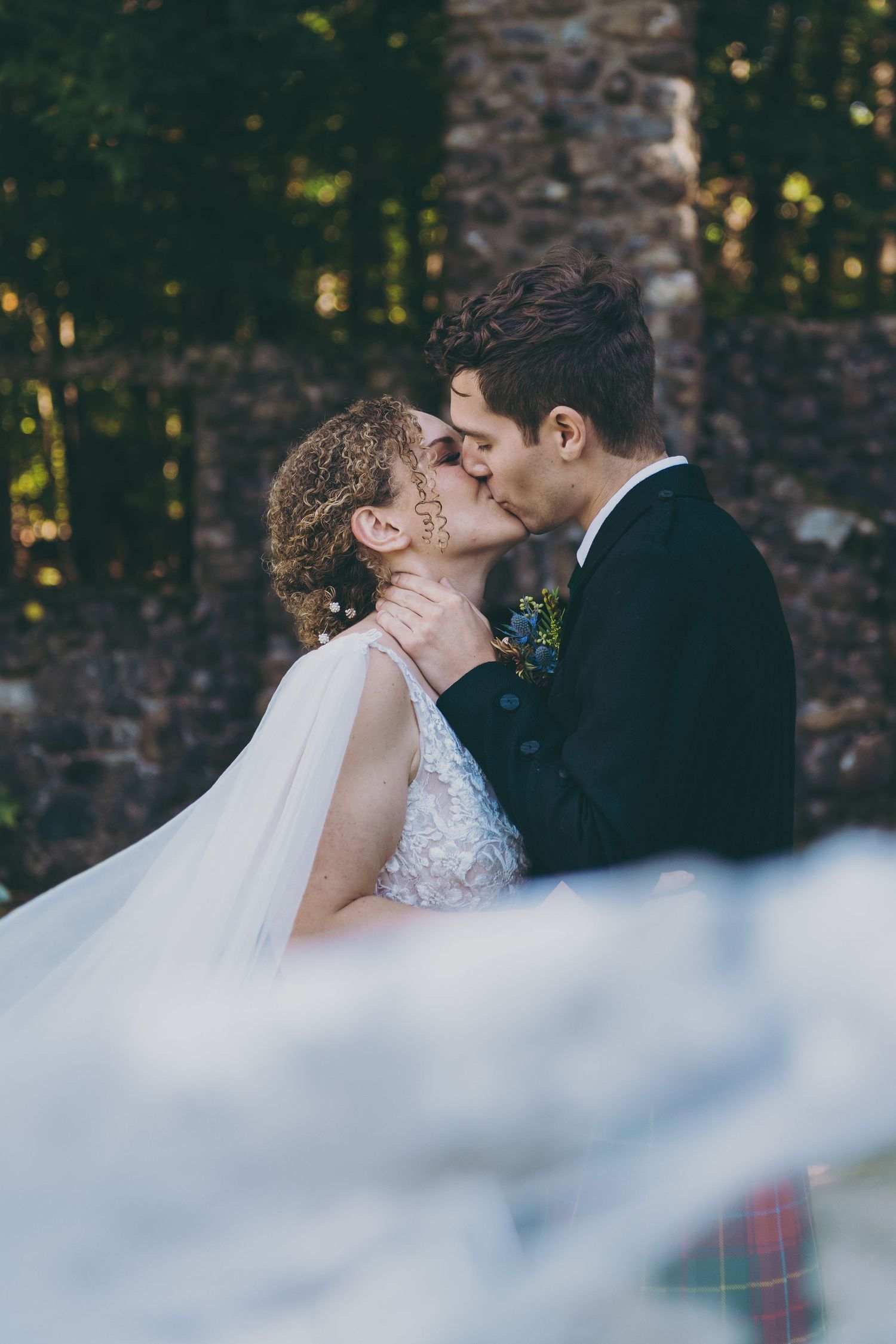 Romantic wedding kiss captured through flowing veil with stone wall backdrop.