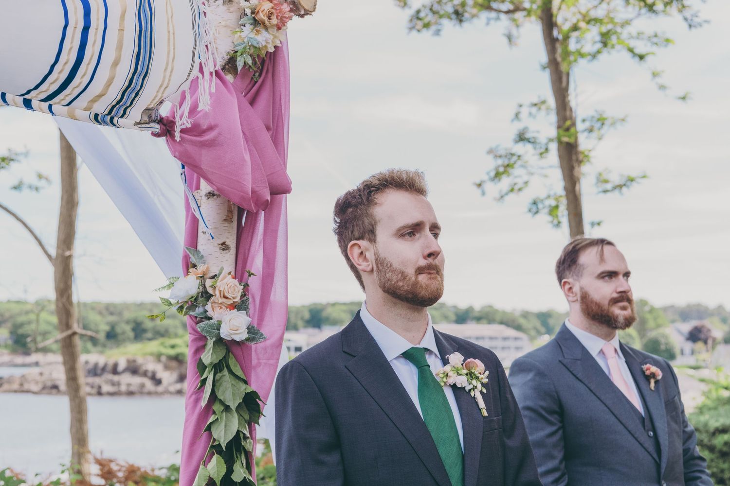 Two wedding attendants in dark suits standing outdoors with a floral-decorated arch and water view in the background.