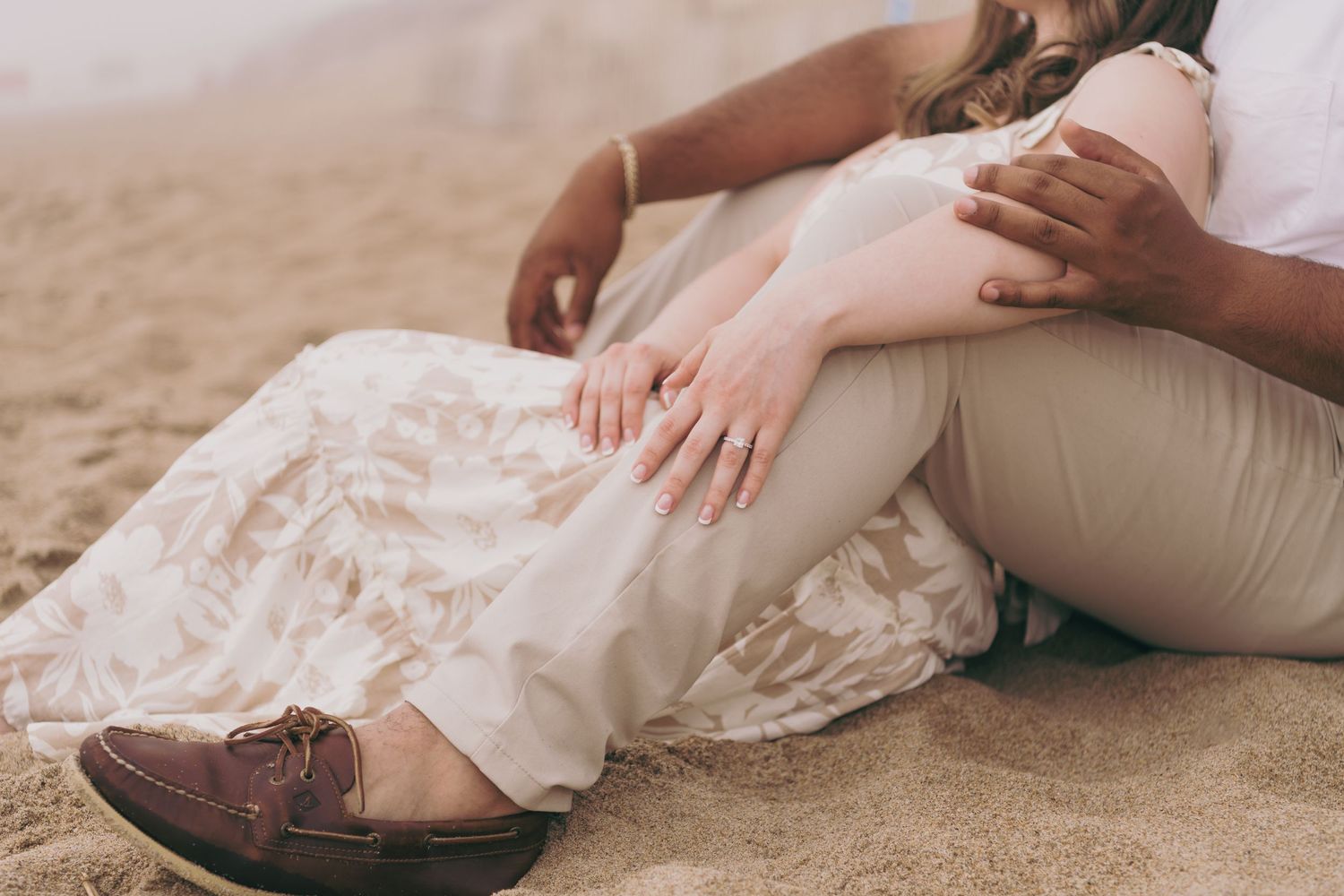 Hands tenderly touch on sandy beach while wearing elegant wedding attire with soft lighting.