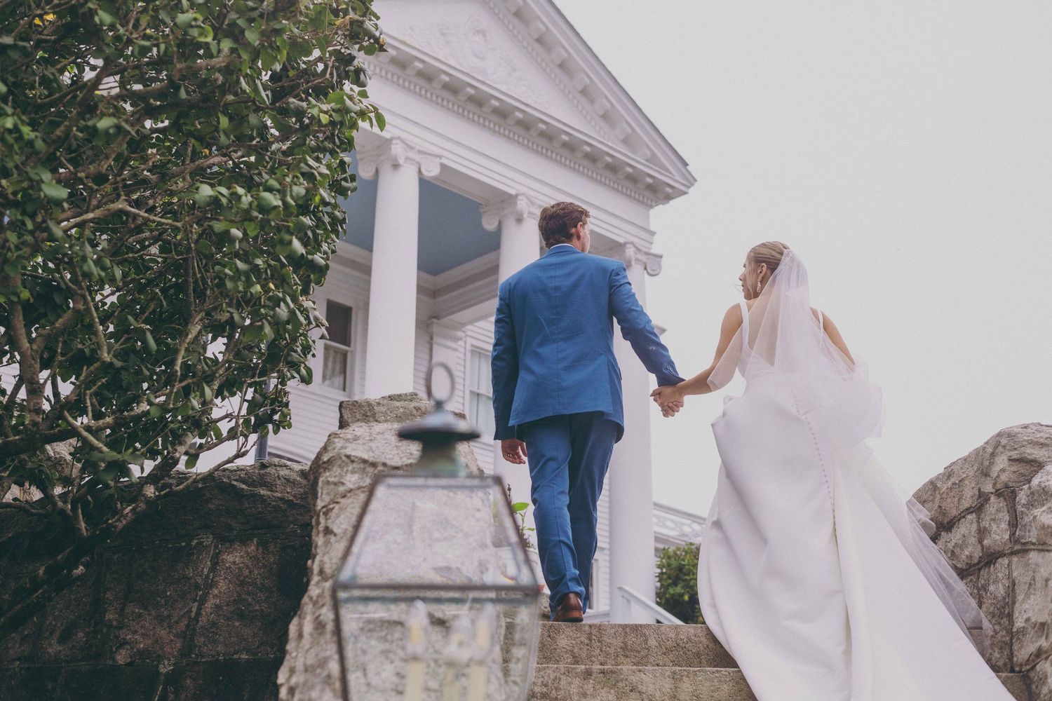 A couple in formal wedding attire walking away from the camera up white stone steps towards a historic building.