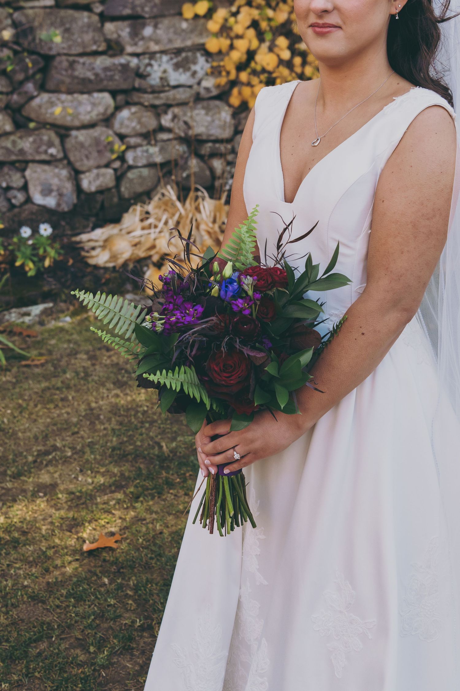 Close-up of bridal bouquet featuring deep purple flowers and green ferns held against white wedding dress.