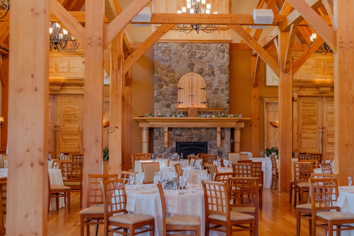 Elegant rustic wedding reception venue with timber beams, stone fireplace and white table settings in warm lighting.