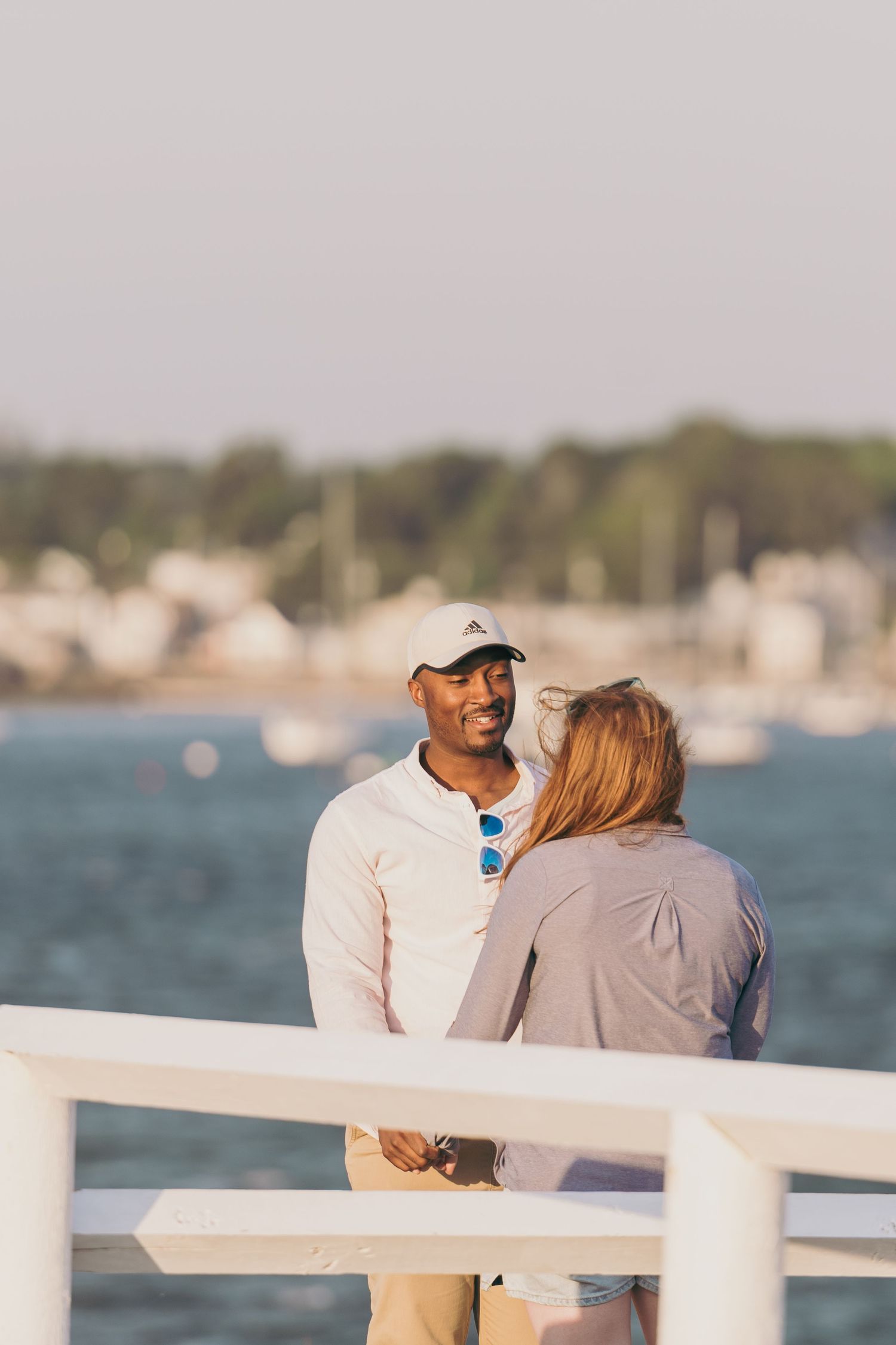 A couple shares a romantic moment while standing at a waterfront railing with boats and harbor views in the background.