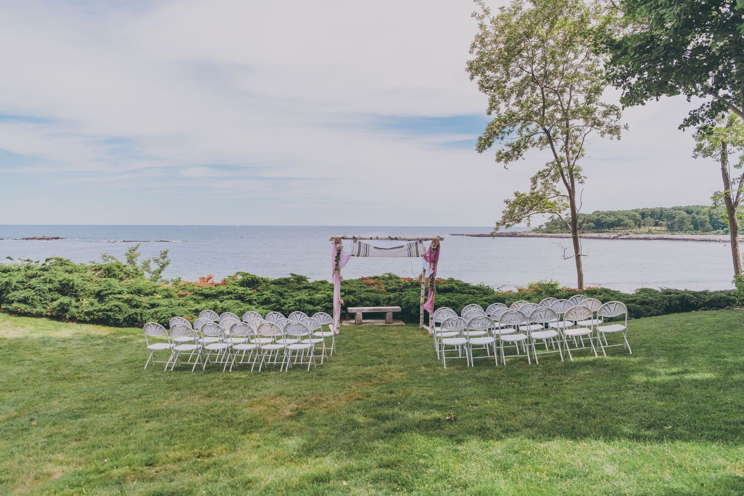 White chairs arranged for an outdoor wedding ceremony with a wooden arch overlooking the ocean on a grassy coastal lawn.