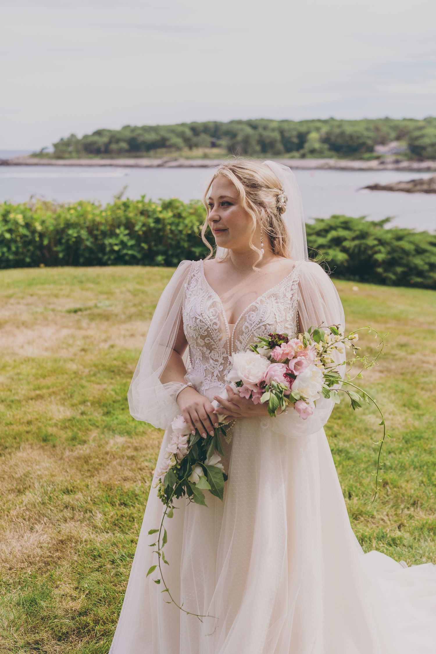 A bride in a flowing white dress with long sleeves holds a pink and white floral bouquet against a coastal landscape background.