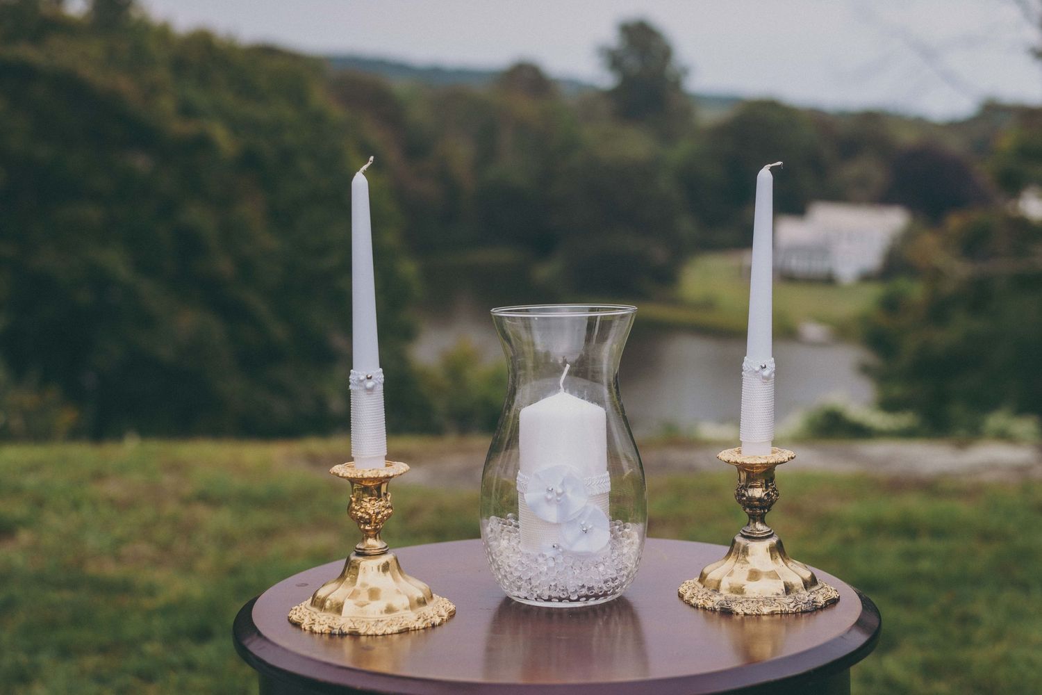 Two lit white candles and a glass vase sit on a wooden surface with a blurred natural outdoor background.