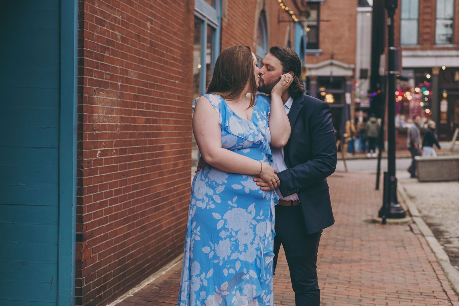 A couple shares a romantic moment on a brick-lined street corner, with one wearing a blue floral dress and the other in black.