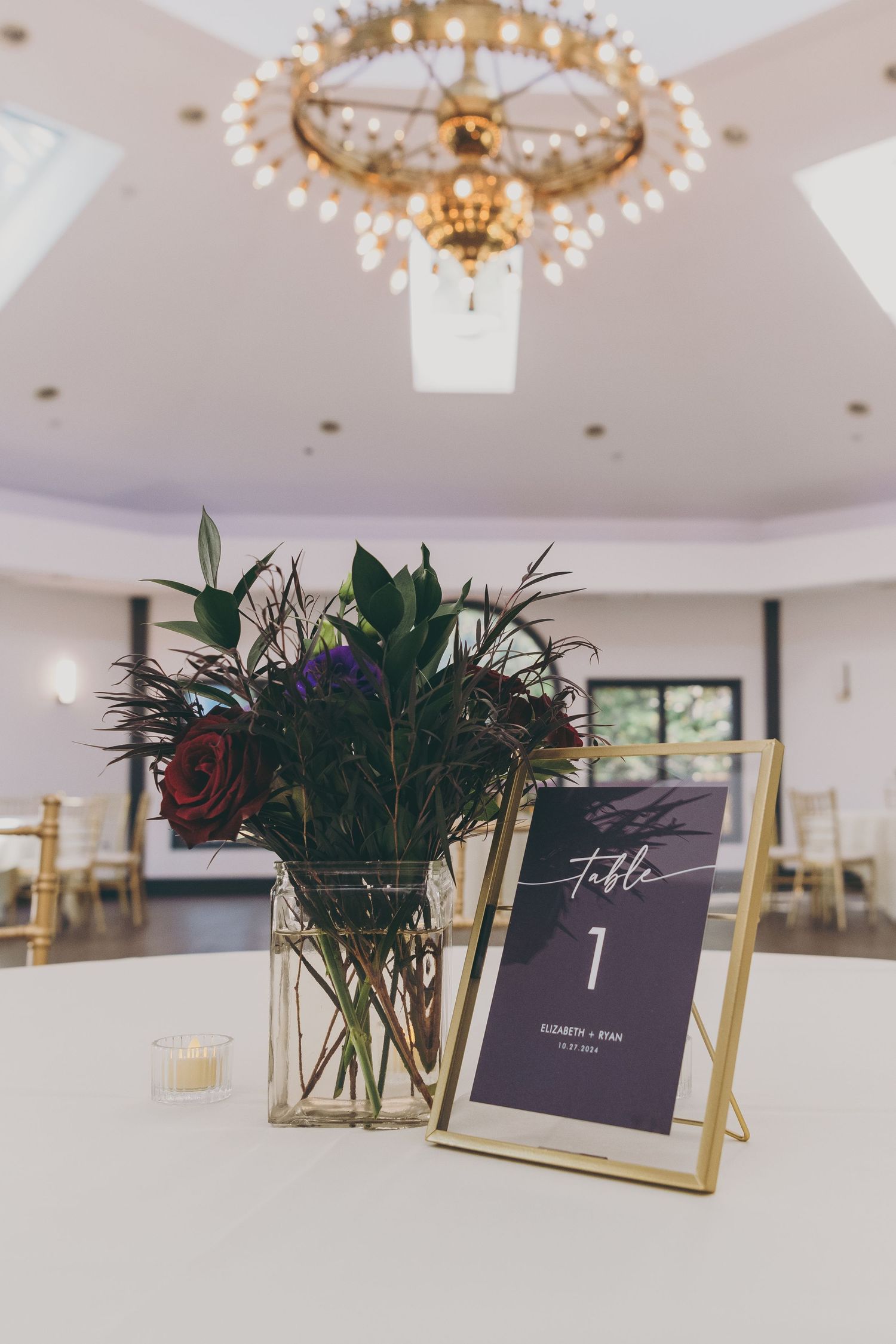 Elegant wedding table numbers in gold frames with dark floral arrangements under crystal chandeliers.