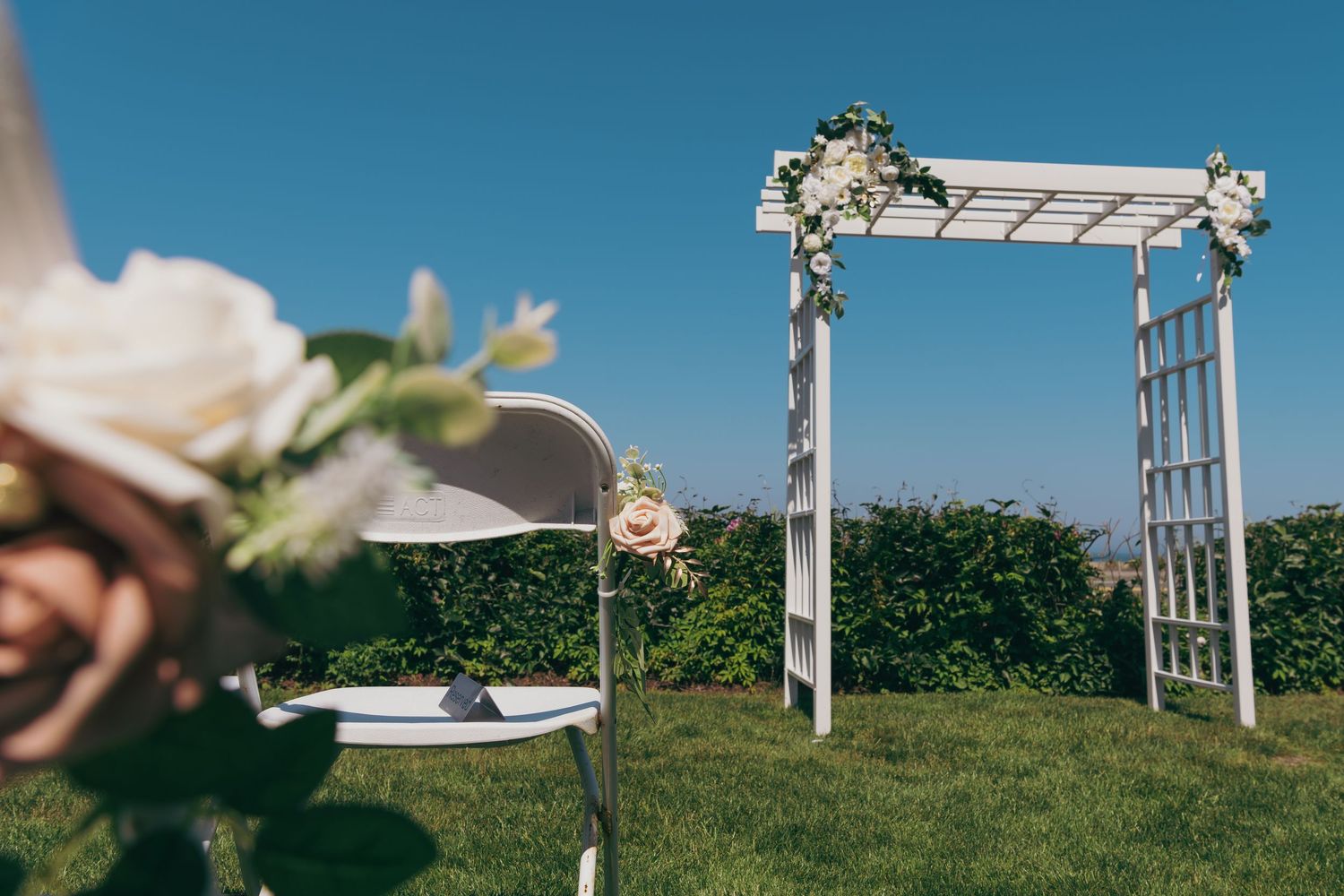 White floral wedding arch stands on a grassy lawn against blue sky with blurred flowers in foreground.