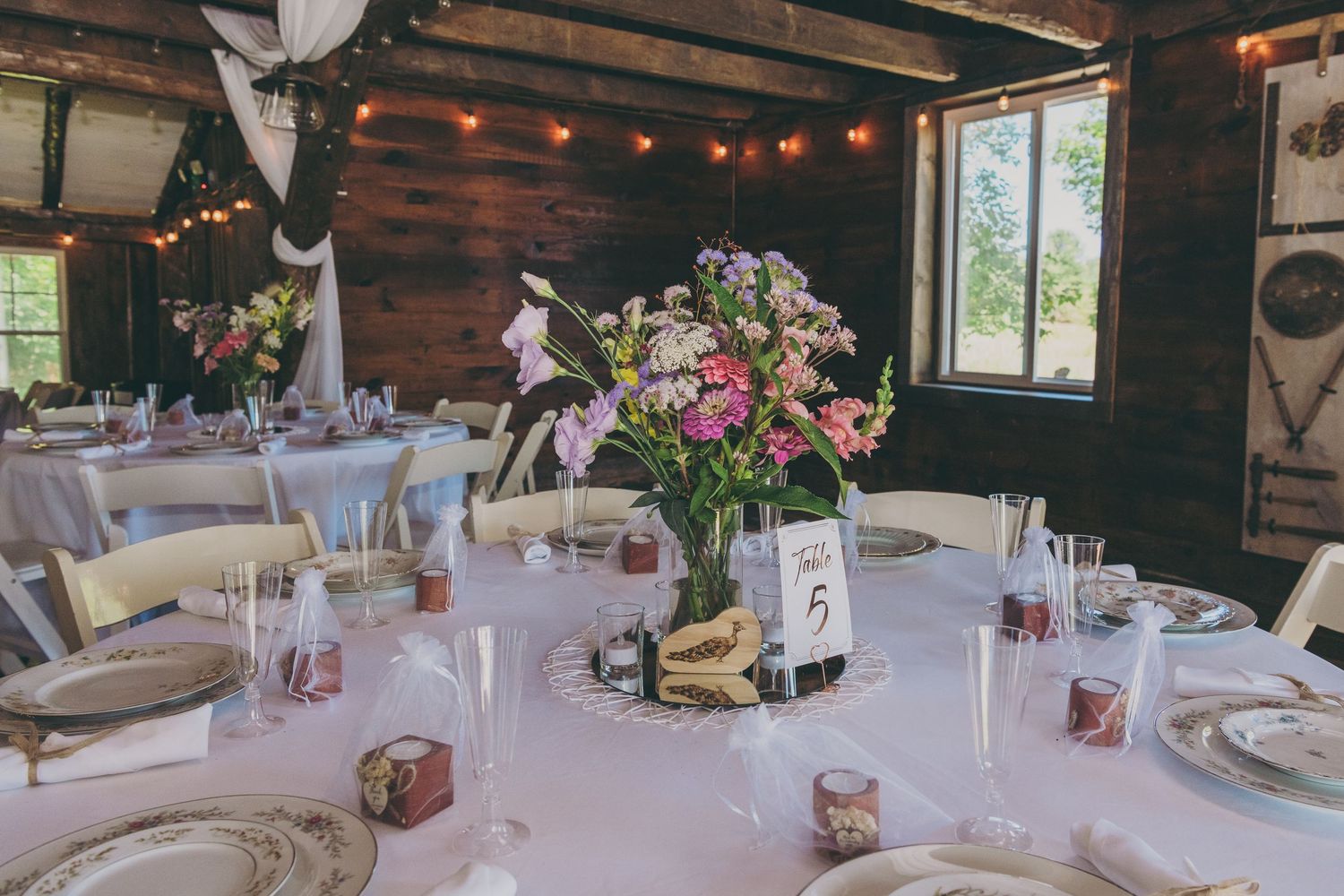 A rustic barn reception setup with white tablecloths, floral centerpieces and string lights hanging from wooden beams.