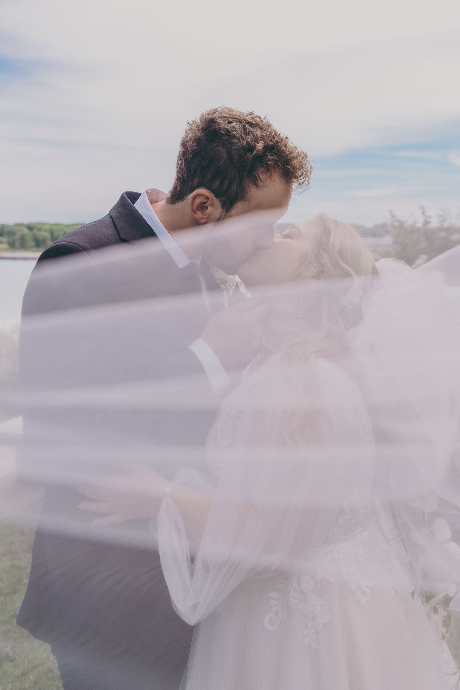 Romantic wedding moment captured through ethereal veil against cloudy sky backdrop.