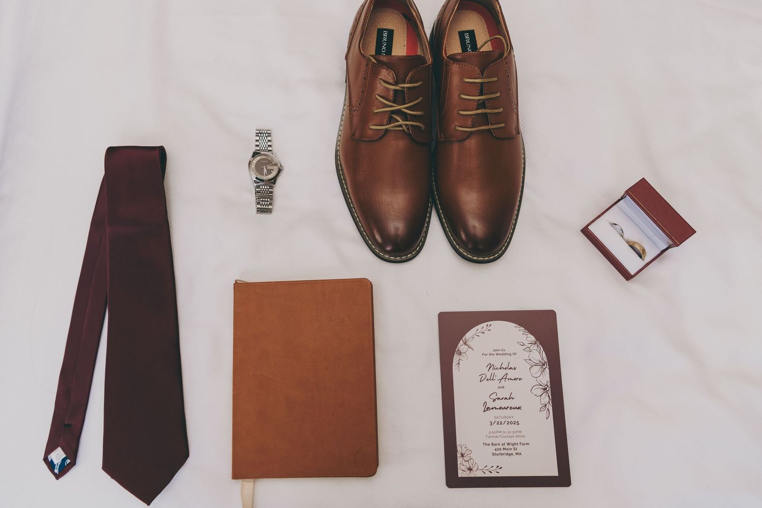 Groom's accessories including brown leather dress shoes, necktie, watch, and wedding invitation on white surface.