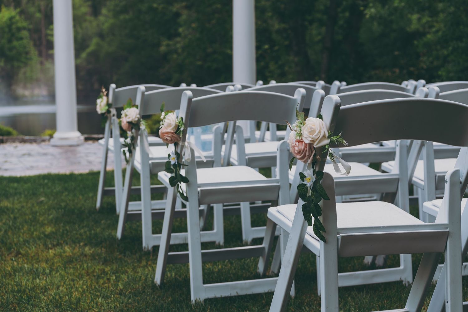 White folding chairs decorated with pink and white floral arrangements line outdoor wedding ceremony rows on green grass.