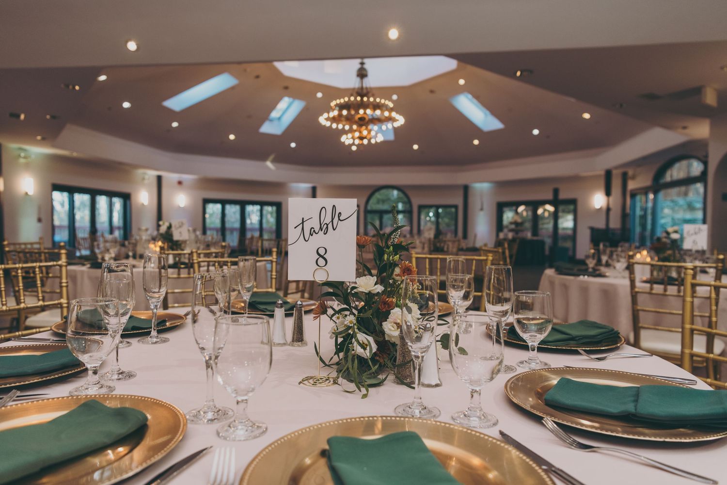 Elegant wedding reception venue with green and gold place settings, round tables, and crystal chandelier lighting.