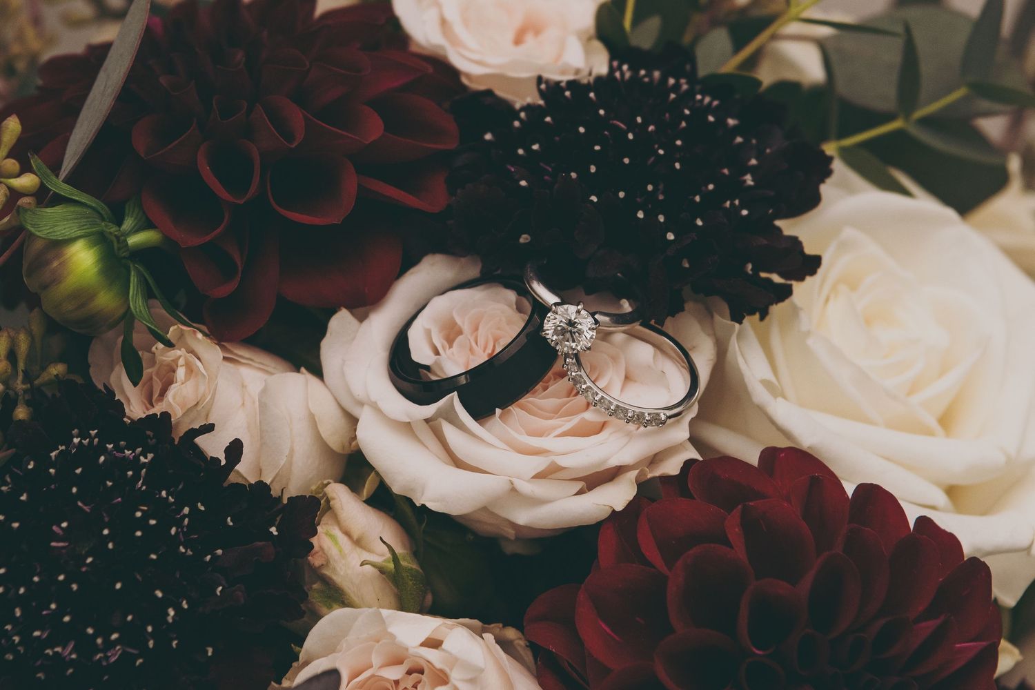 Wedding rings nestled in light pink roses surrounded by deep burgundy dahlias and greenery with a sparkly effect.