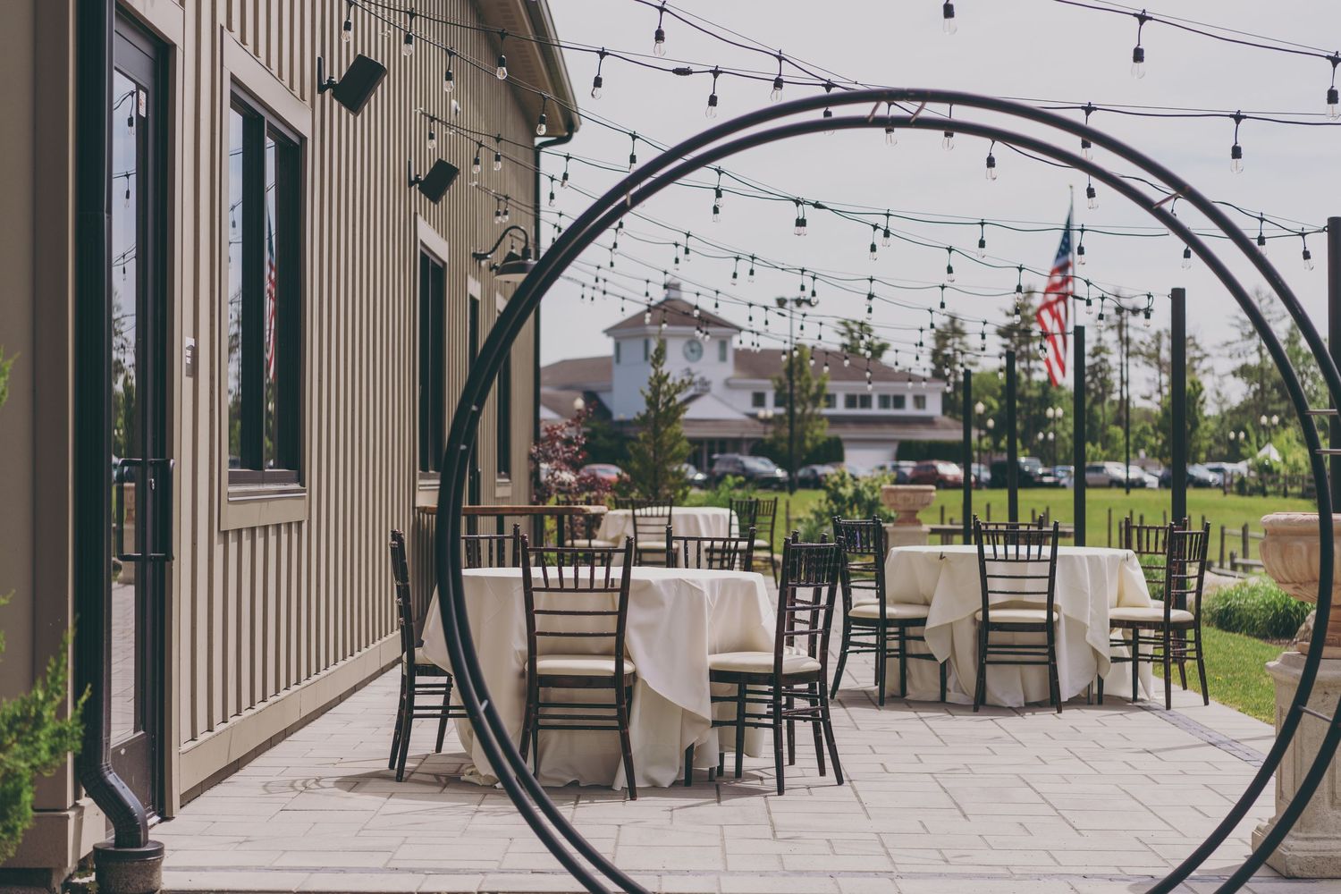 A modern outdoor patio with string lights and dining tables under an arched metal framework, overlooking a waterfront.
