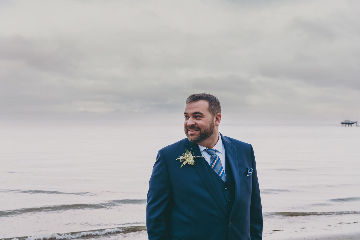 Person in navy three-piece suit smiling while standing near ocean shore on overcast day.