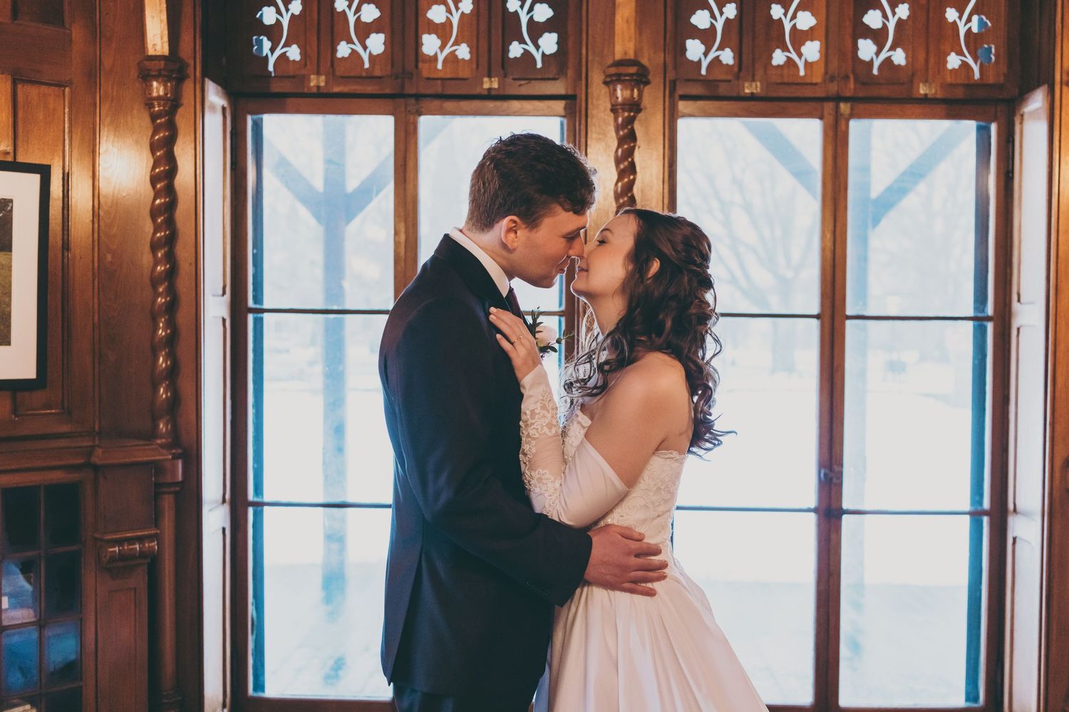 A romantic wedding moment between a couple kissing in front of an ornate window with decorative woodwork.