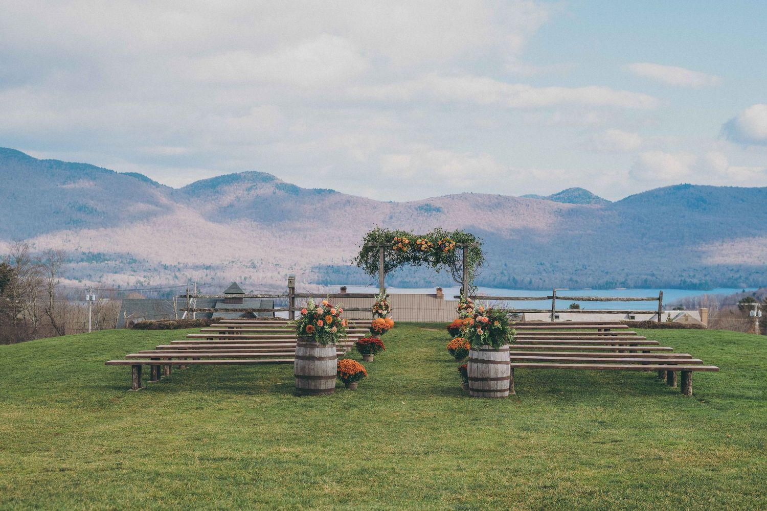 Wedding ceremony setup with wooden benches and floral arch overlooking scenic mountain landscape on grassy hilltop.