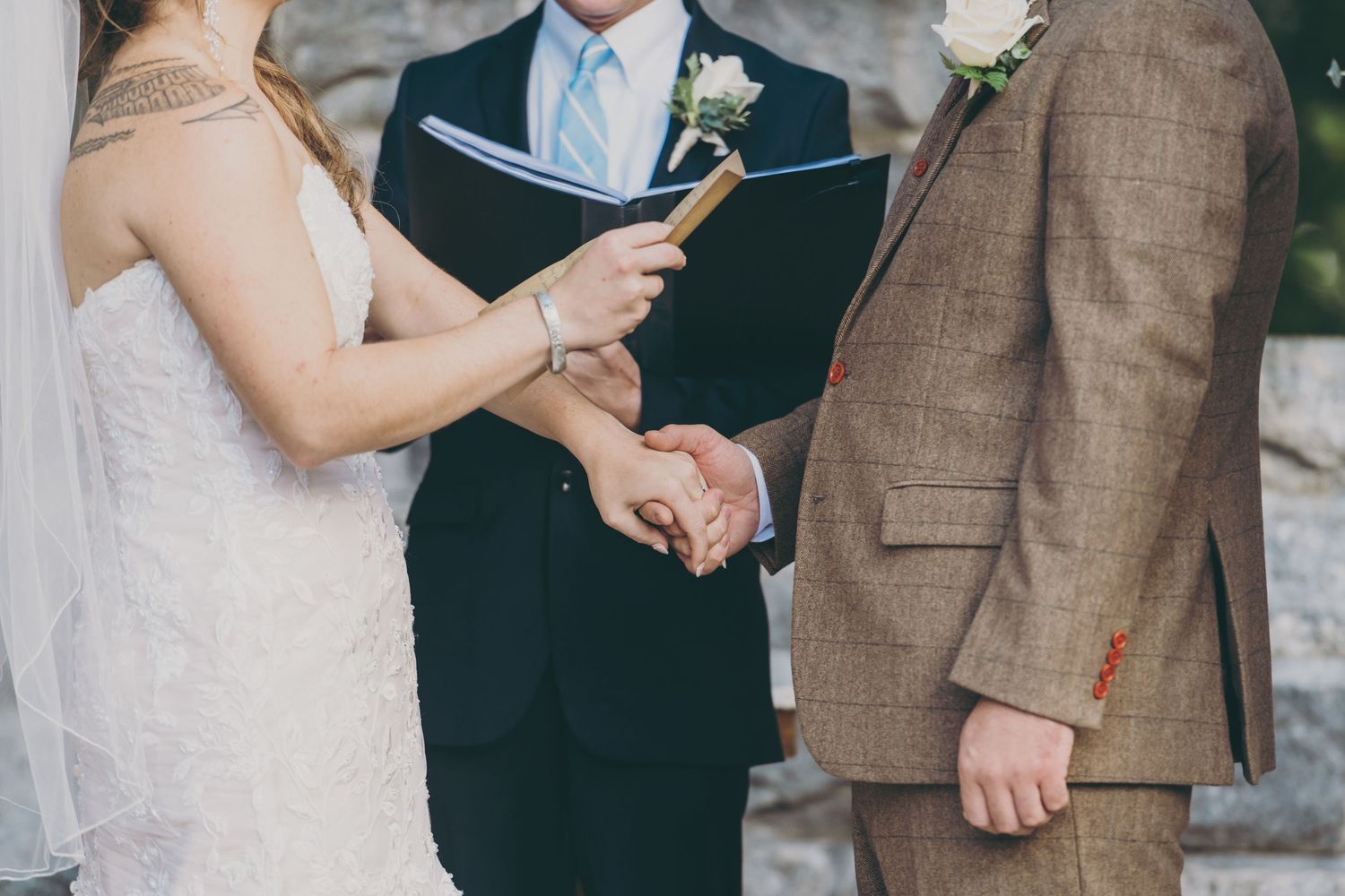 A couple holds hands during an outdoor wedding ceremony while the officiant reads from a book.
