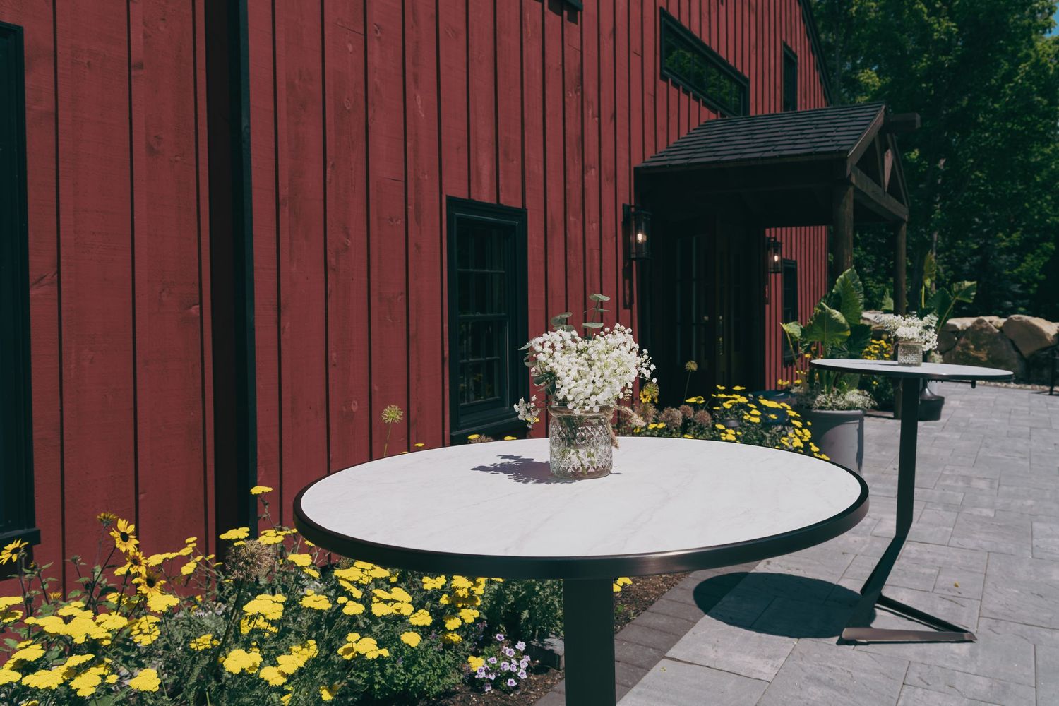 A white garden table with chairs surrounded by yellow flowers and a red barn-style building in the background.