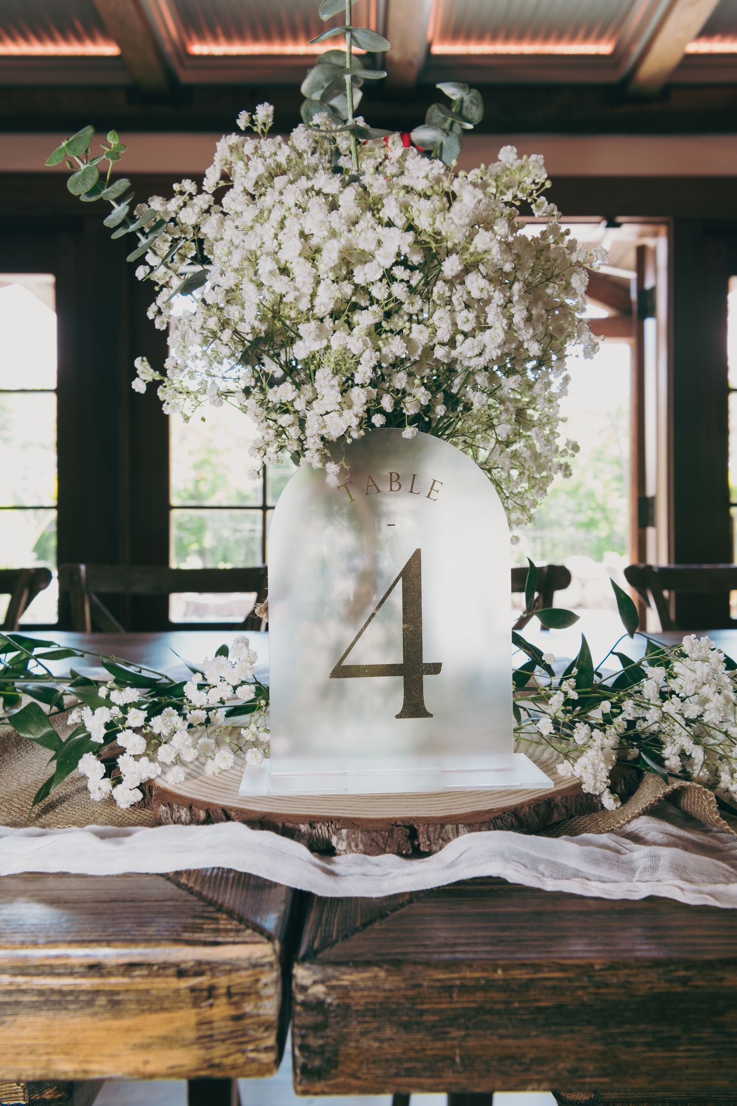 Elegant wedding table centerpiece with white baby's breath flowers in a vintage milk jug with number 4 on rustic wooden surface.