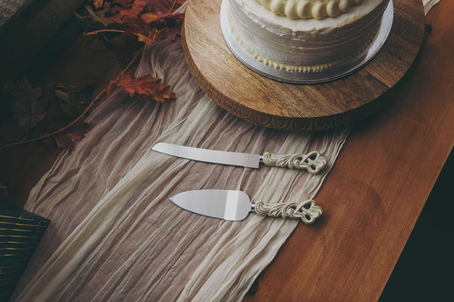 Ornate silver cake serving utensils rest on a gray linen napkin beside a wooden plate and cake stand.