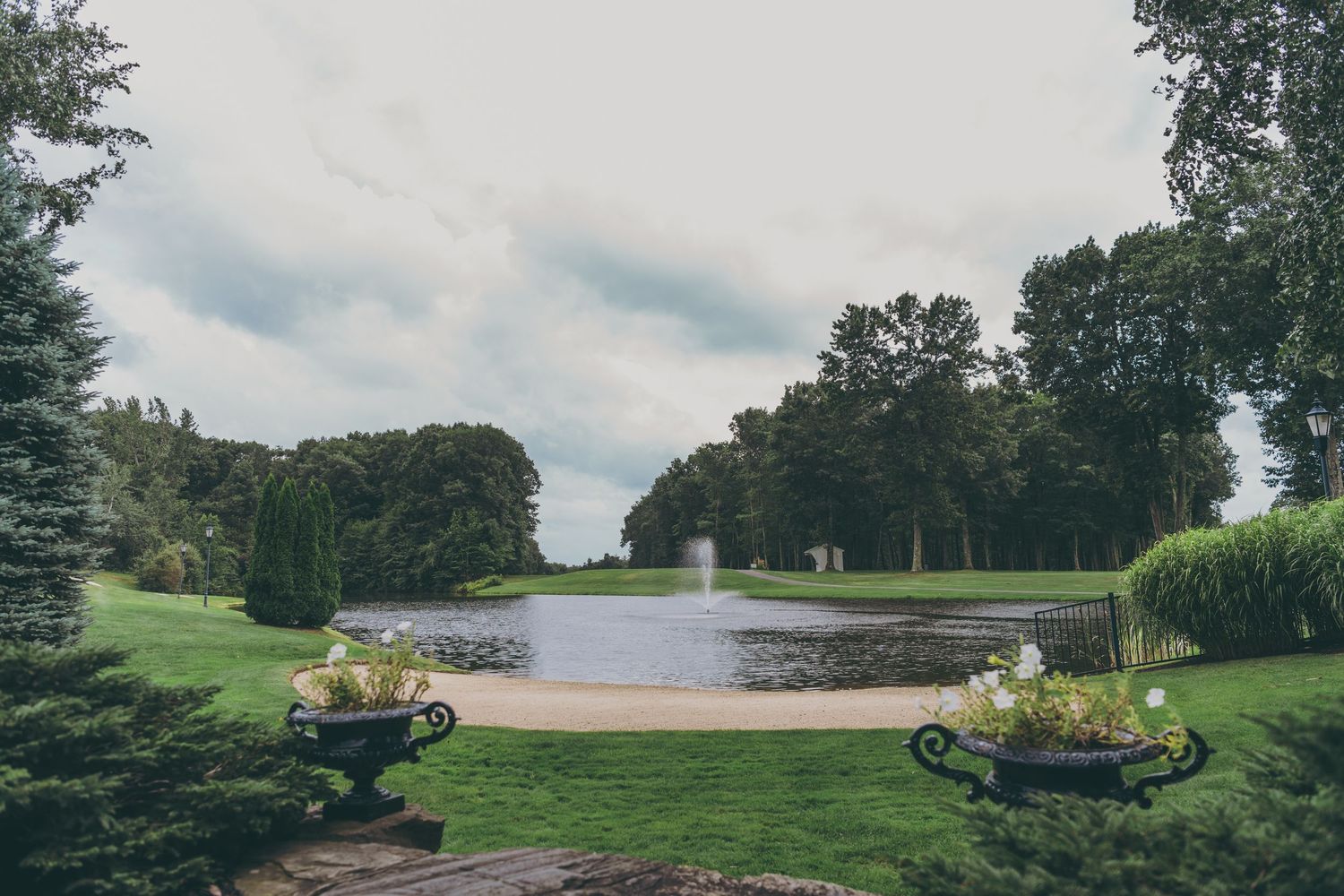 An elegant pond surrounded by manicured lawn and ornamental gardens with decorative urns on a cloudy summer day.