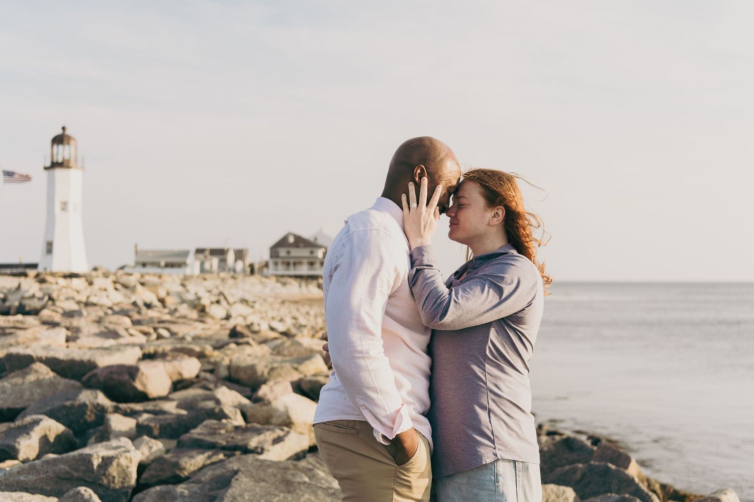 A romantic couple embraces near a lighthouse on a rocky coastal beach at sunset.
