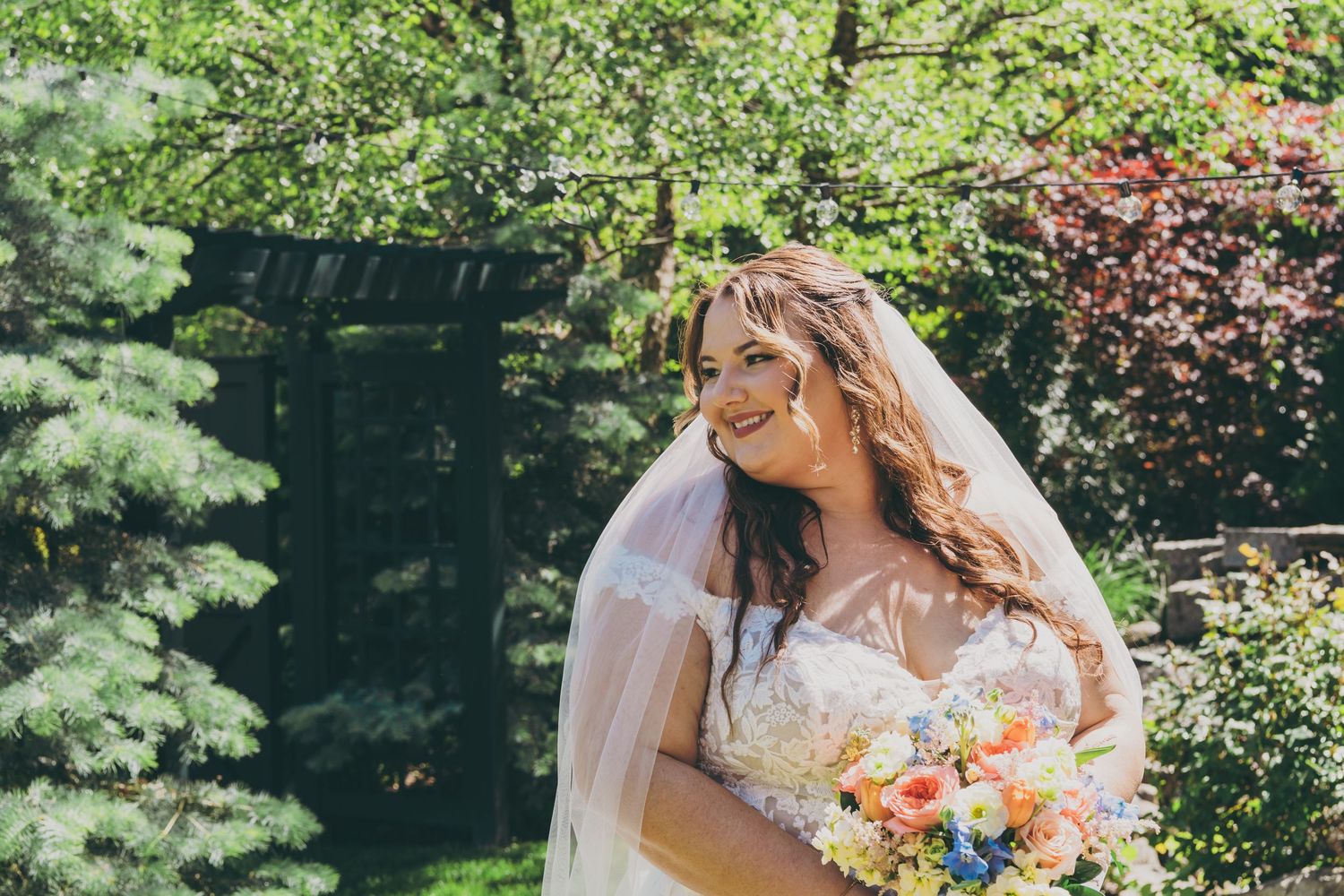 Bride in lace wedding dress holds colorful peach and coral bouquet in sunlit garden setting.