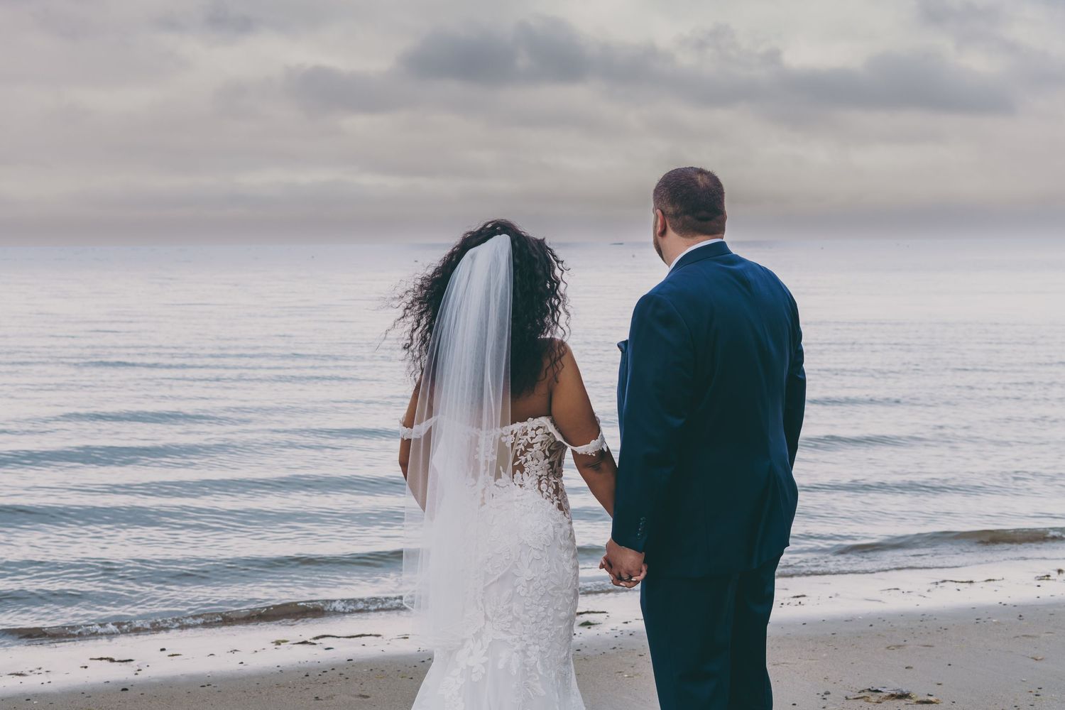 A newlywed couple stands at the ocean's edge during an overcast beach wedding ceremony.