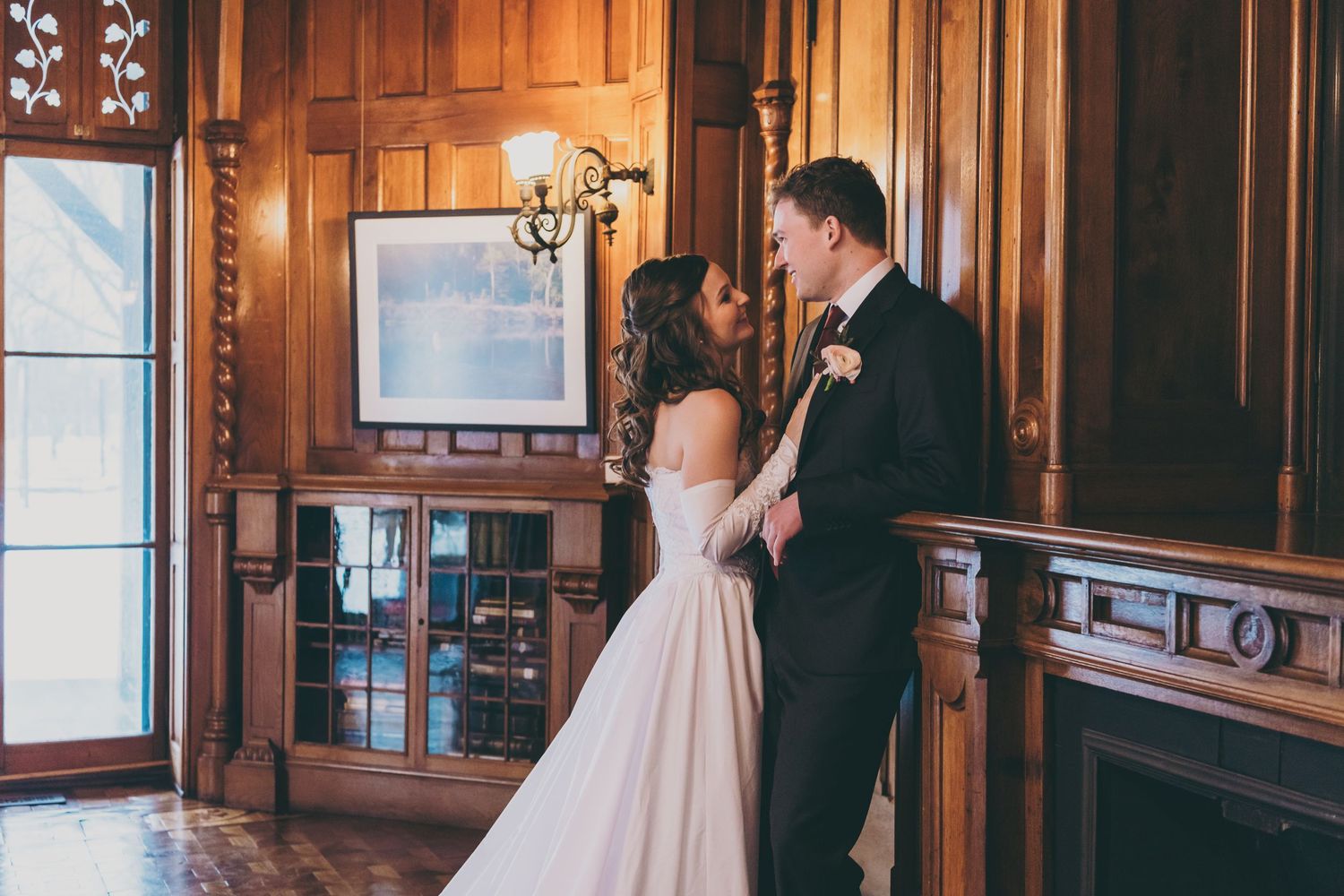 A tender embrace between newlyweds in an elegant wood-paneled room with natural light streaming through windows.