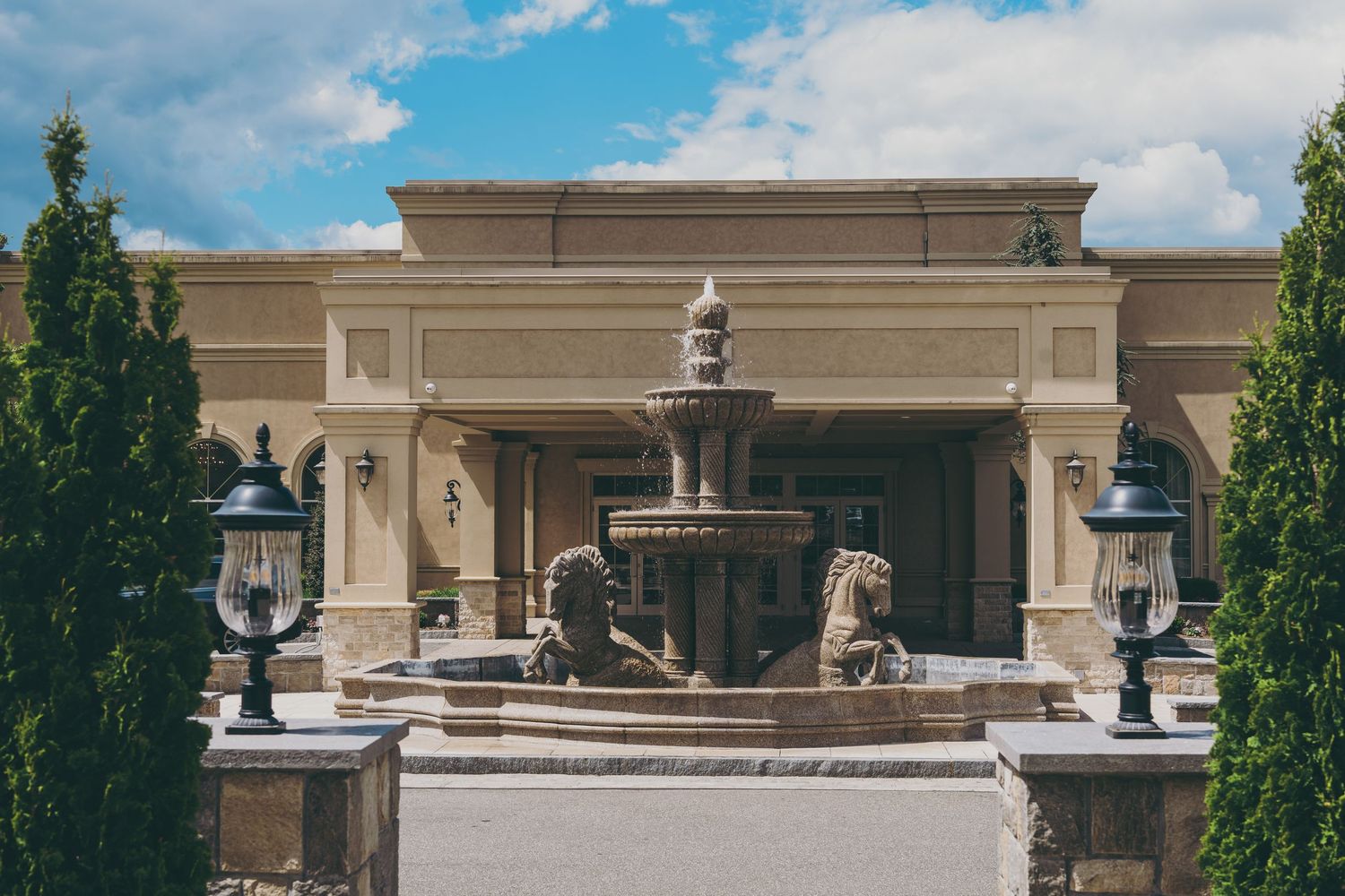 Elegant stone fountain with ornate details stands in front of a classical beige building entrance with decorative lamps.