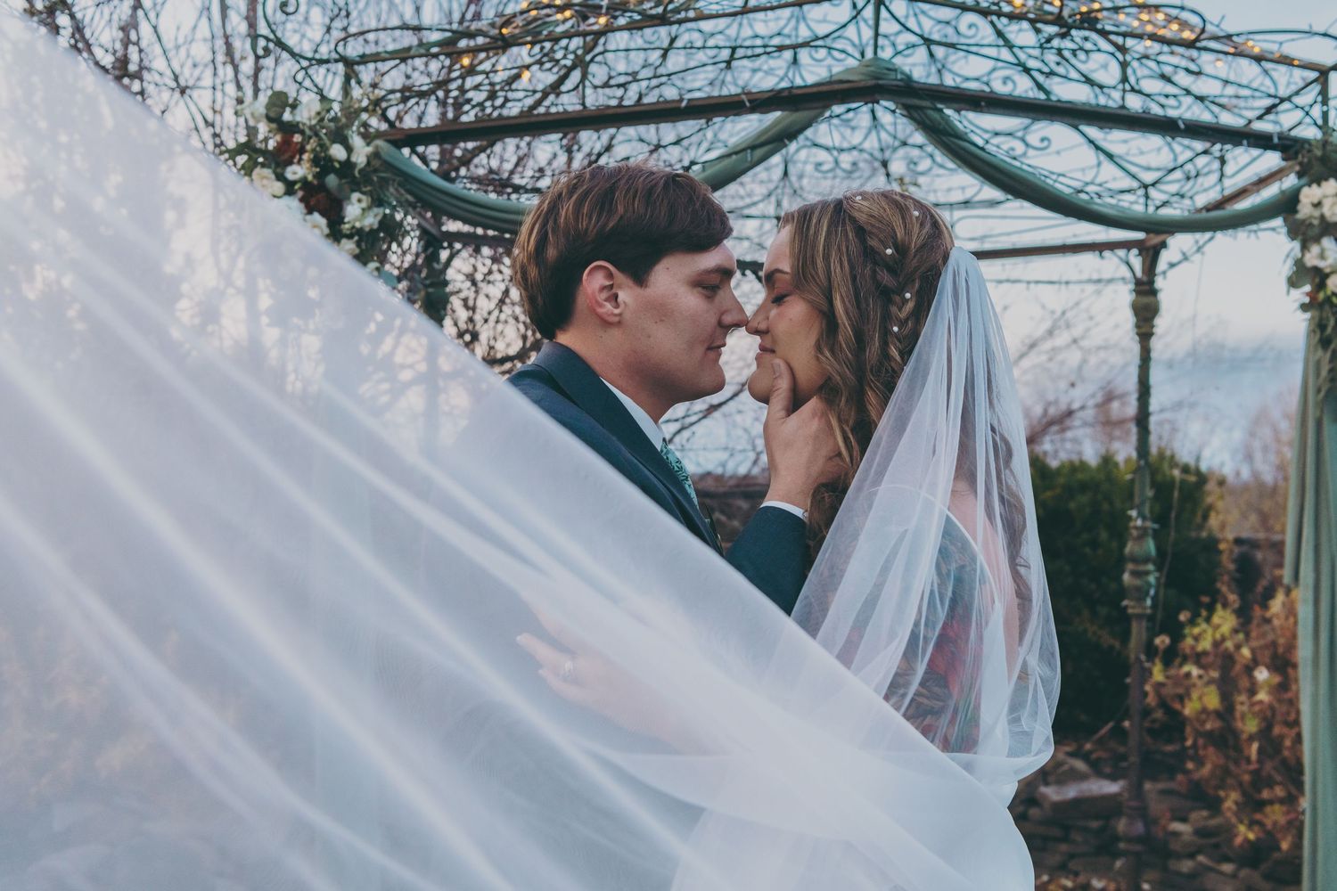 A romantic wedding moment captured under a decorative garden gazebo with flowing white veil in the breeze.