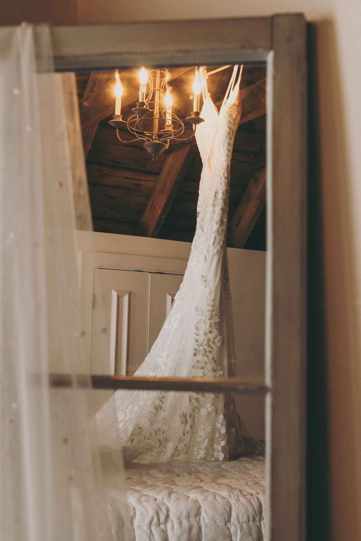 A delicate white lace wedding dress hangs in a dimly lit room with a glowing chandelier reflection in the mirror.