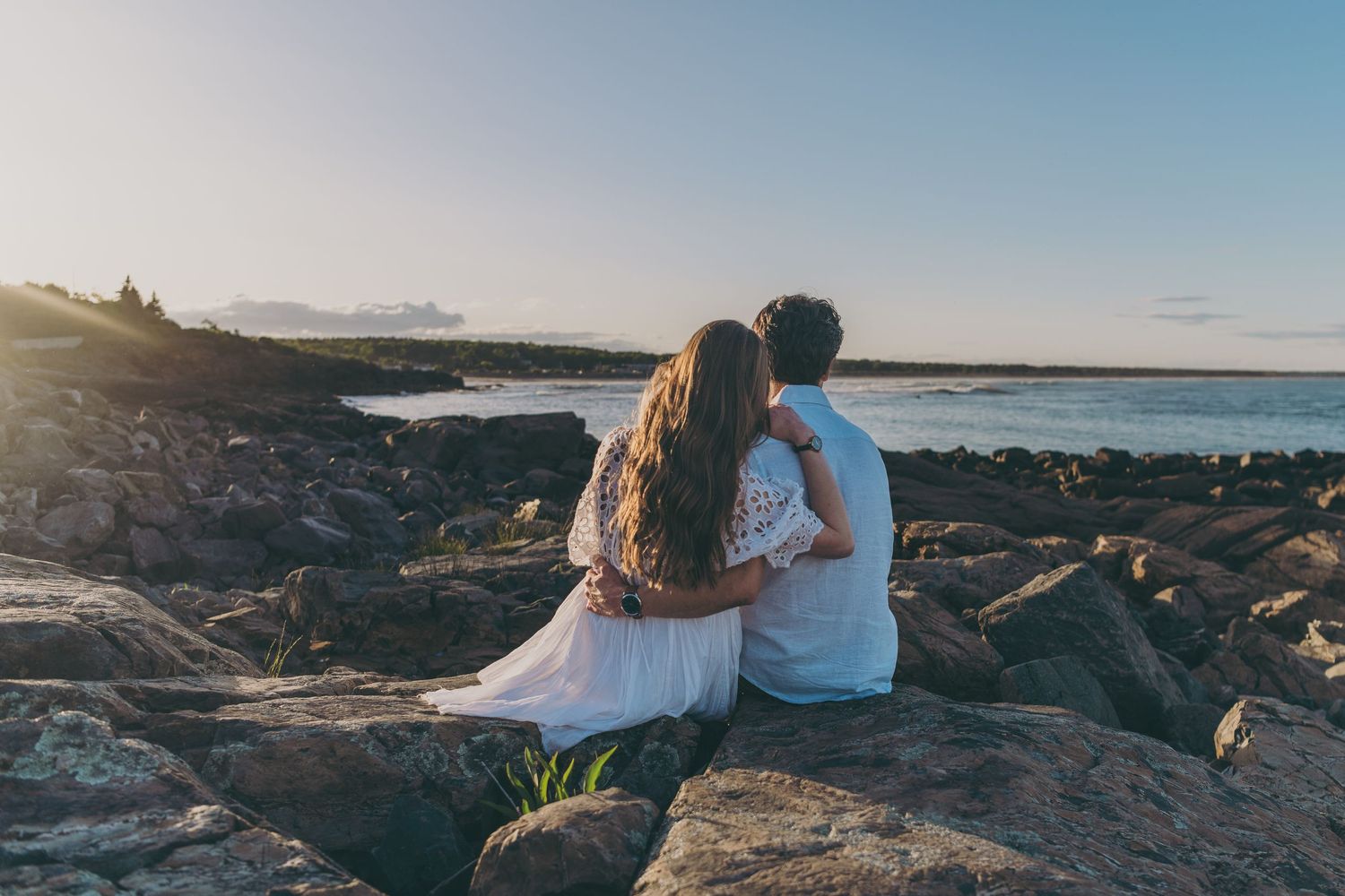 Couple sits together on rocky oceanside cliff during sunset watching the waves crash against the shore.