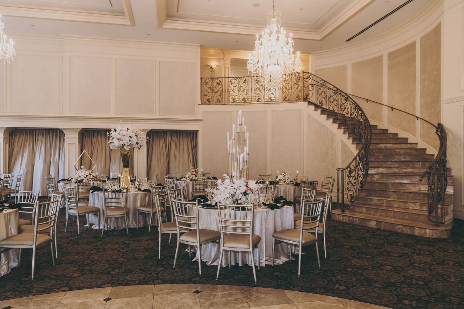 Elegant ballroom with crystal chandeliers, grand curved staircase, and round tables set for a formal wedding reception.