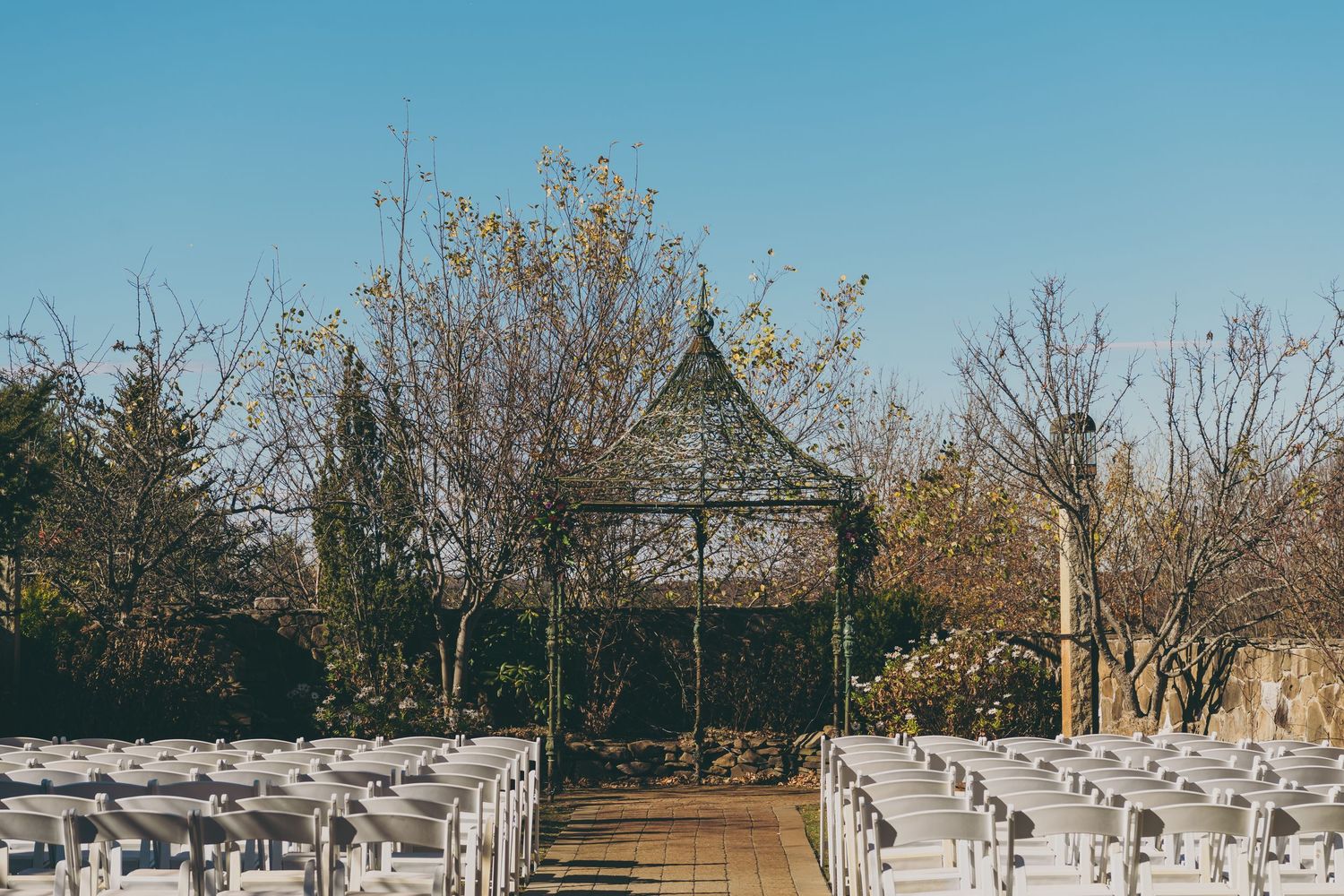 White folding chairs line a garden path leading to a rustic wooden gazebo surrounded by bare trees in an outdoor venue setup.