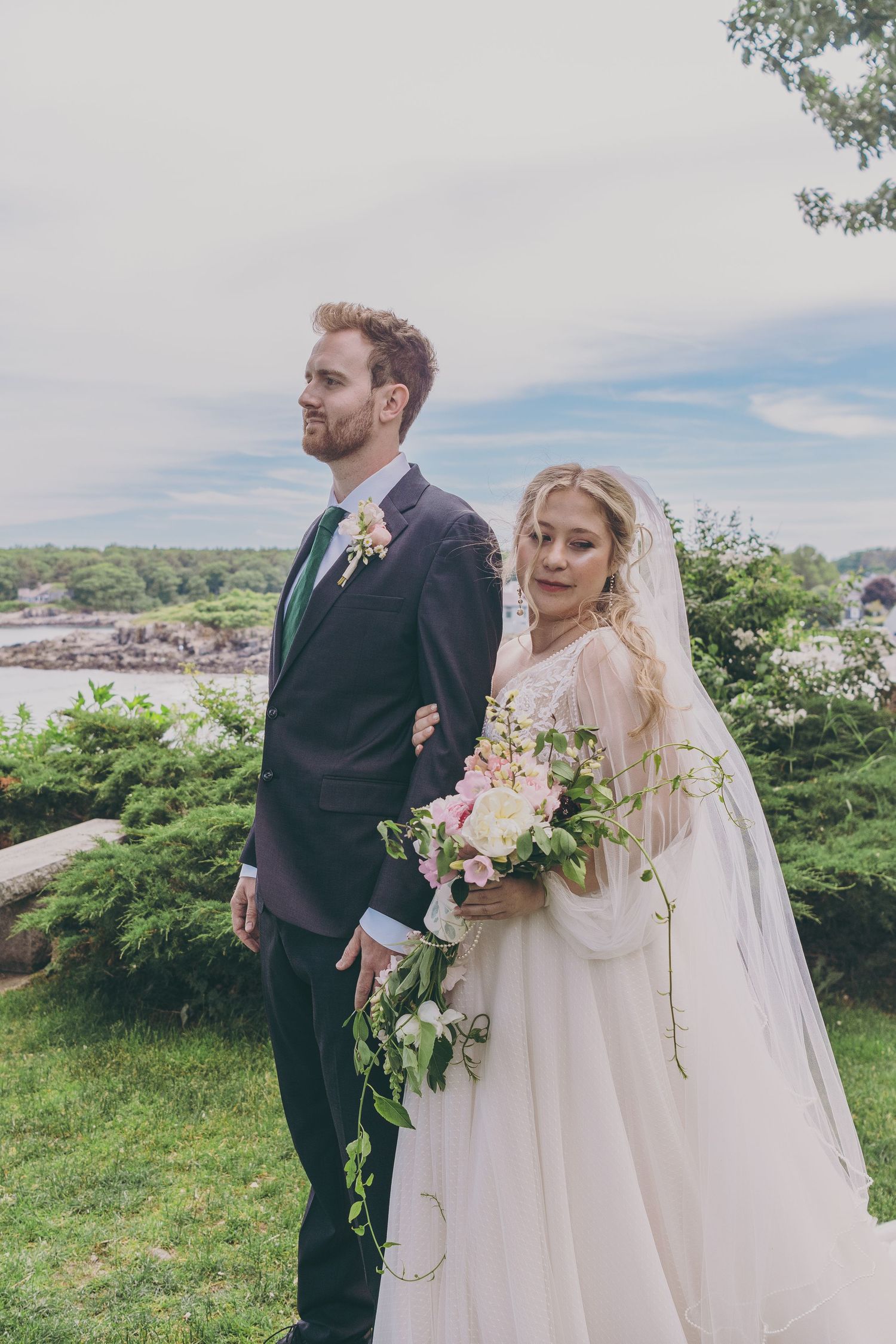 Newlyweds pose outdoors with scenic coastal backdrop and flowing bridal bouquet.