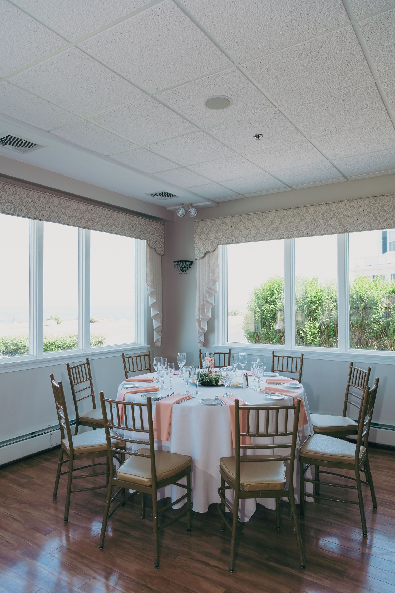 A bright dining space with panoramic windows features round white tablecloths and dark wooden chairs in a row.