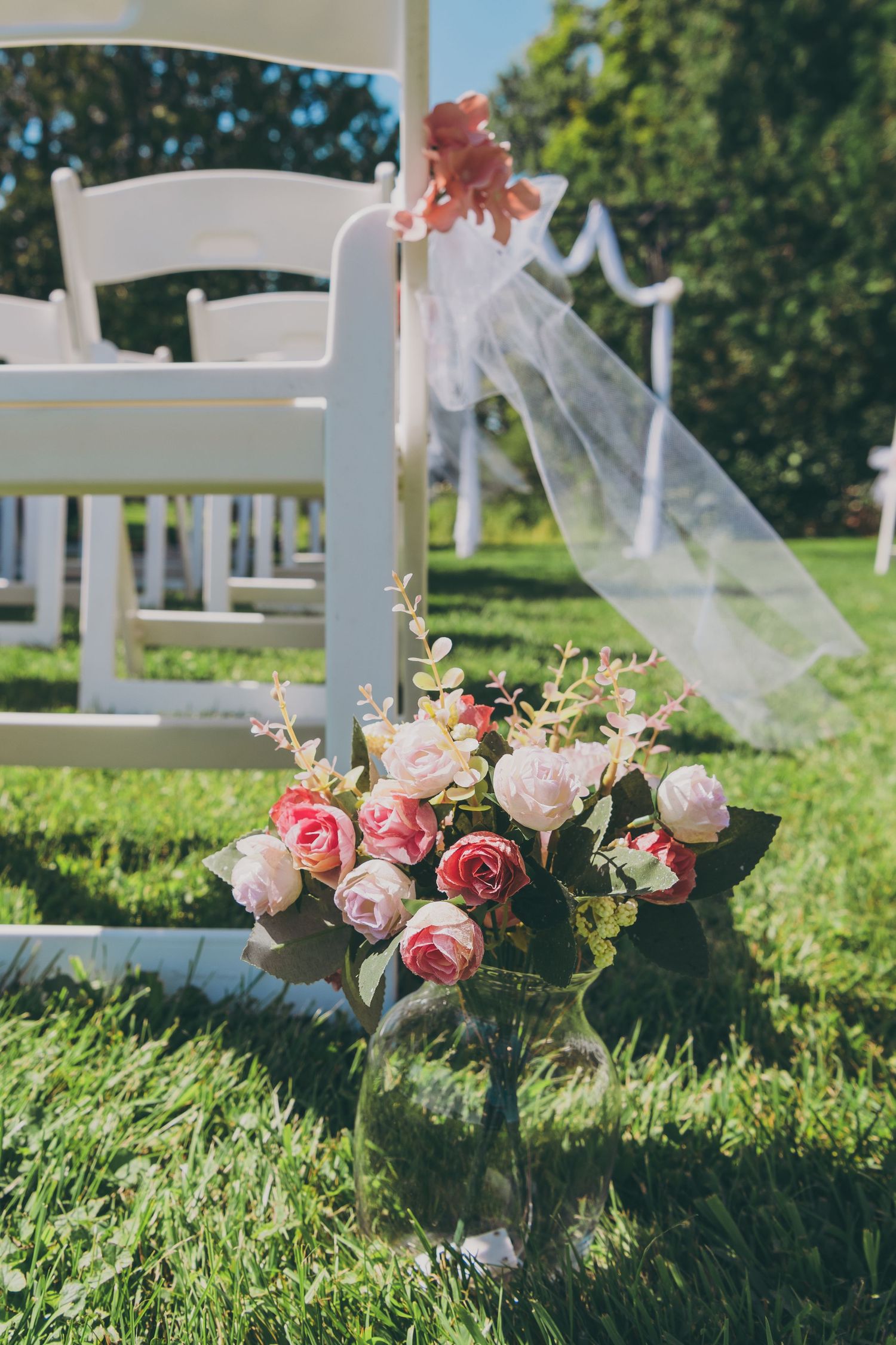 White wedding ceremony chair decorated with pink roses and flowing tulle ribbon in an outdoor garden setting.
