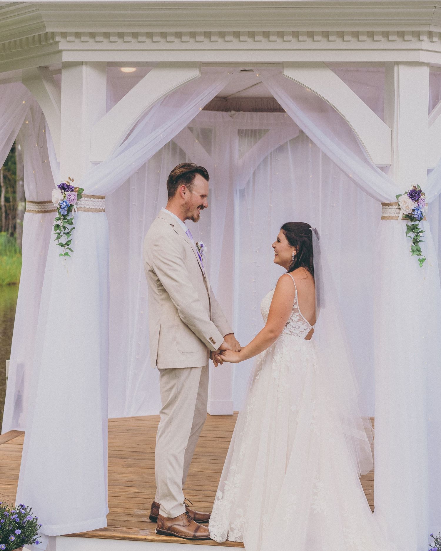 A bride and groom hold hands under a white draped wedding gazebo decorated with purple flowers.
