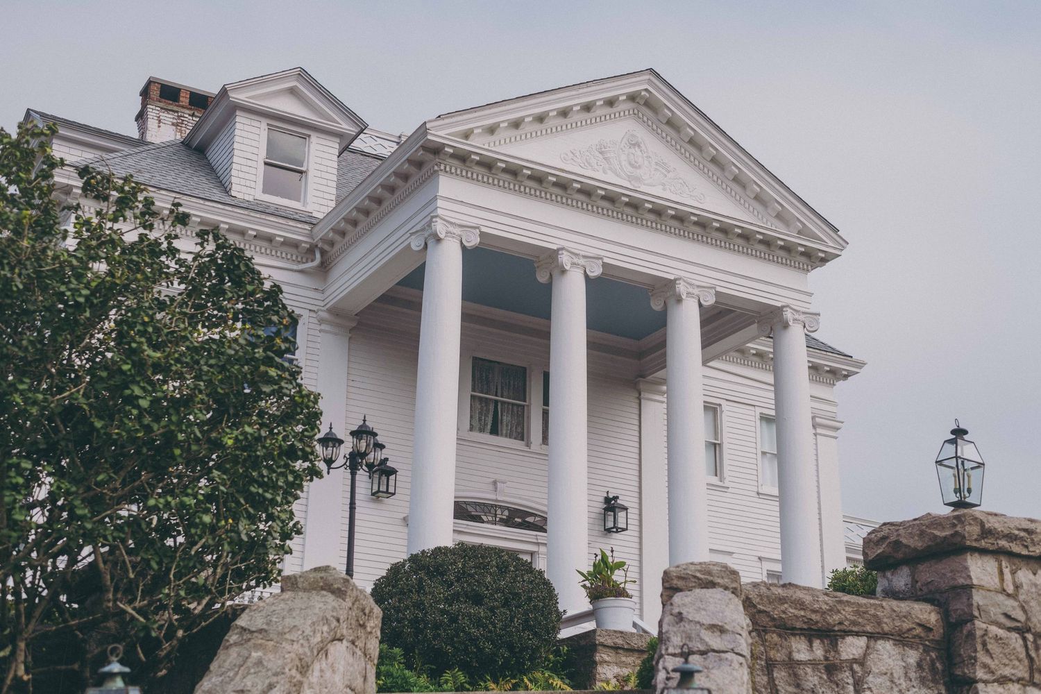 Elegant white Southern mansion with grand columns and ornate architectural details features a triangular pediment and stone wall.