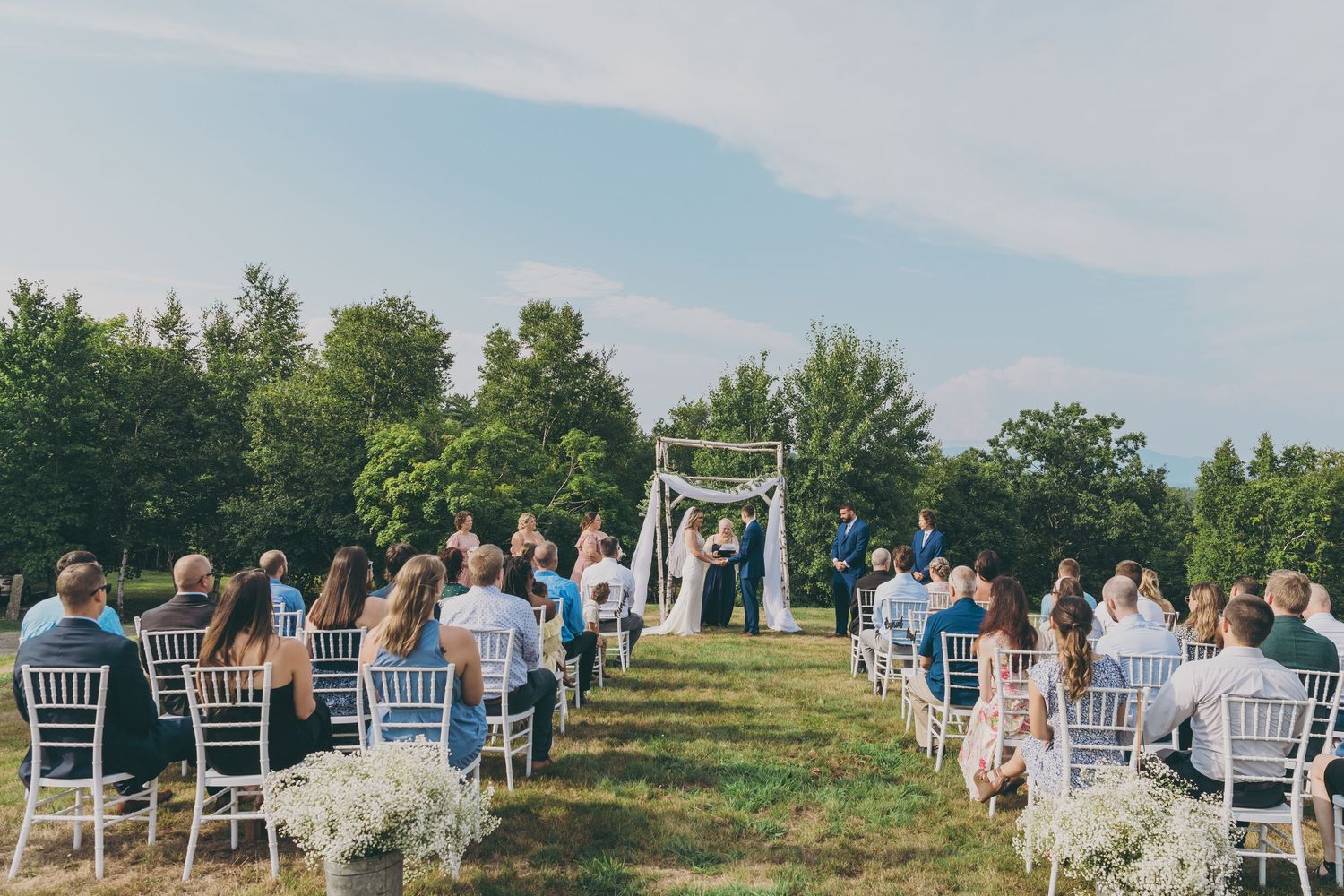 Outdoor wedding ceremony setup with white chairs and floral arch against blue sky.