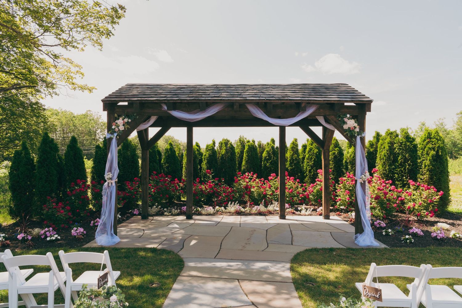 Wedding ceremony gazebo decorated with white fabric and flowers in a garden setting with chairs.