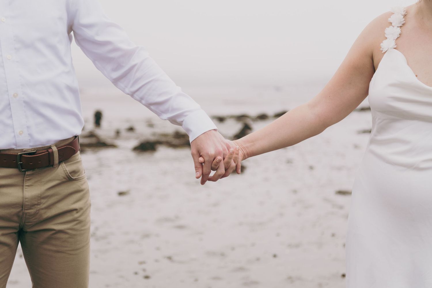 A close-up shot of two people holding hands on a sandy beach, wearing white and beige wedding attire.